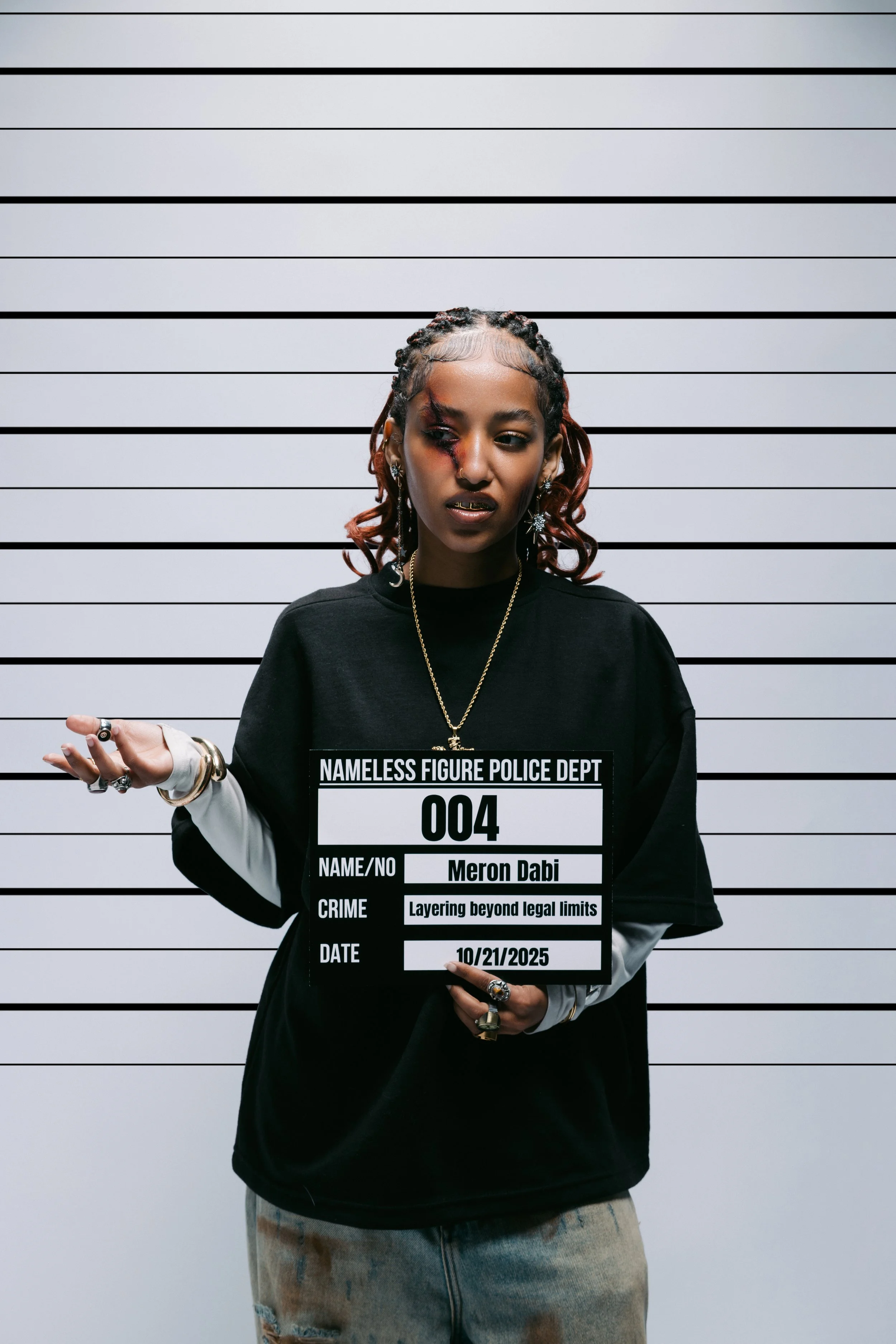 A young woman with braided and styled hair, wearing jewelry, holding a mock police mugshot sign with her name and details, standing in front of a height measurement wall.