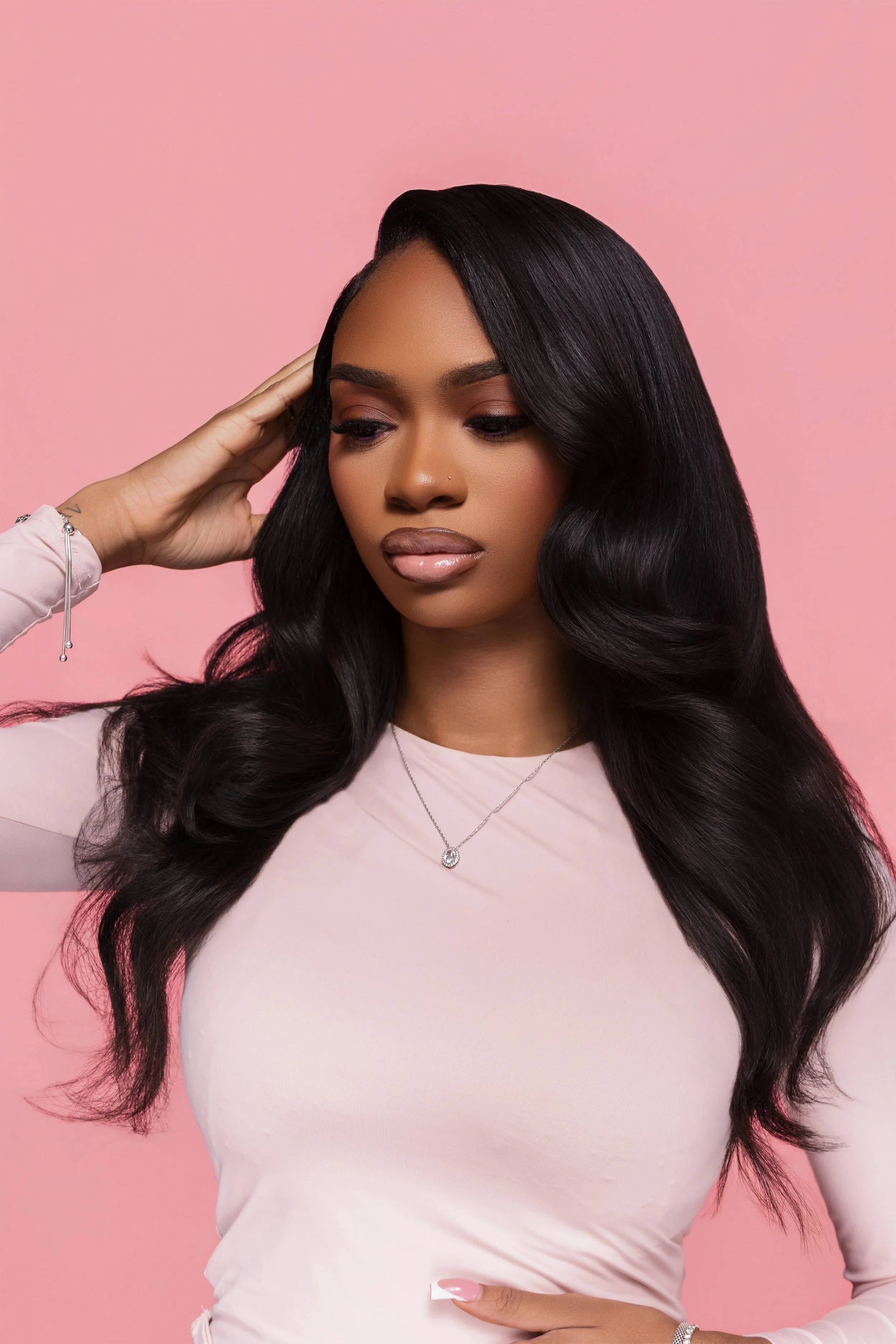 A woman with long, wavy black hair wearing a light pink long-sleeve shirt, a diamond necklace, and a silver bracelet, standing against a pink background.