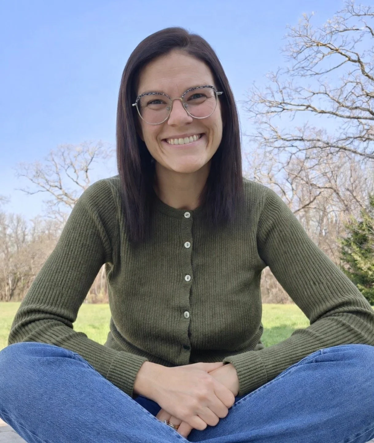 A woman with dark hair and glasses smiling outdoors, sitting cross-legged on grass with trees in the background.