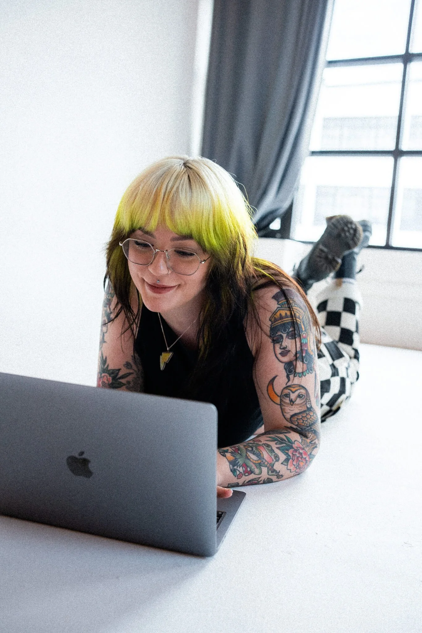 Marci, brand and web designer has green and black hair, glasses, and tattoos on her arms, is leaning over a laptop and smiling. She is indoors with large windows and curtains in the background.