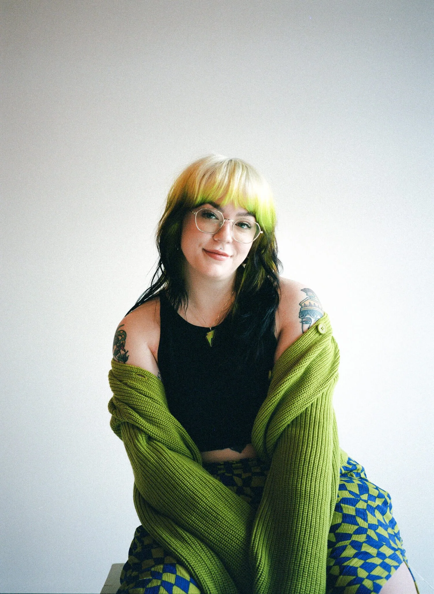 Marci, brand and web designer has green and black hair, and glasses, is sitting in front of a plain white wall, with a bright green cardigan draped over her shoulders and a blue and green checkerboard skirt.