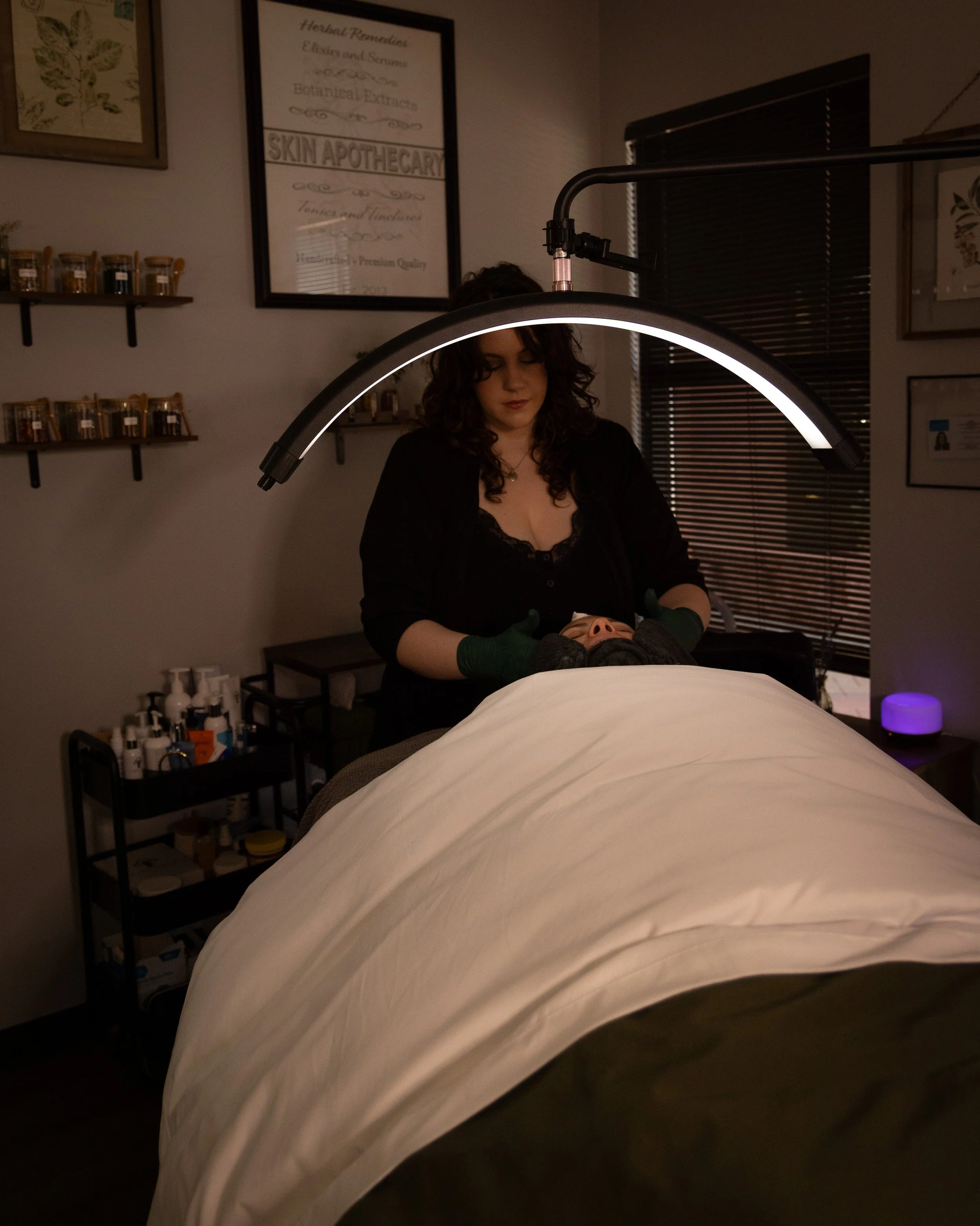 A woman in a black top performing a facial treatment or massage on a client lying face-up on a treatment table, illuminated by an adjustable light, in a spa or skincare clinic setting.