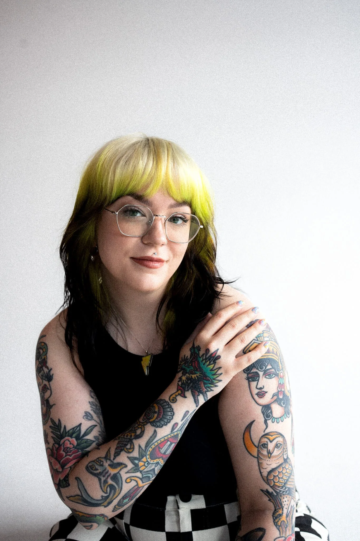 Marci, brand and web designer with green and black hair, glasses, and tattoos on her arms is sitting against a plain white background.
