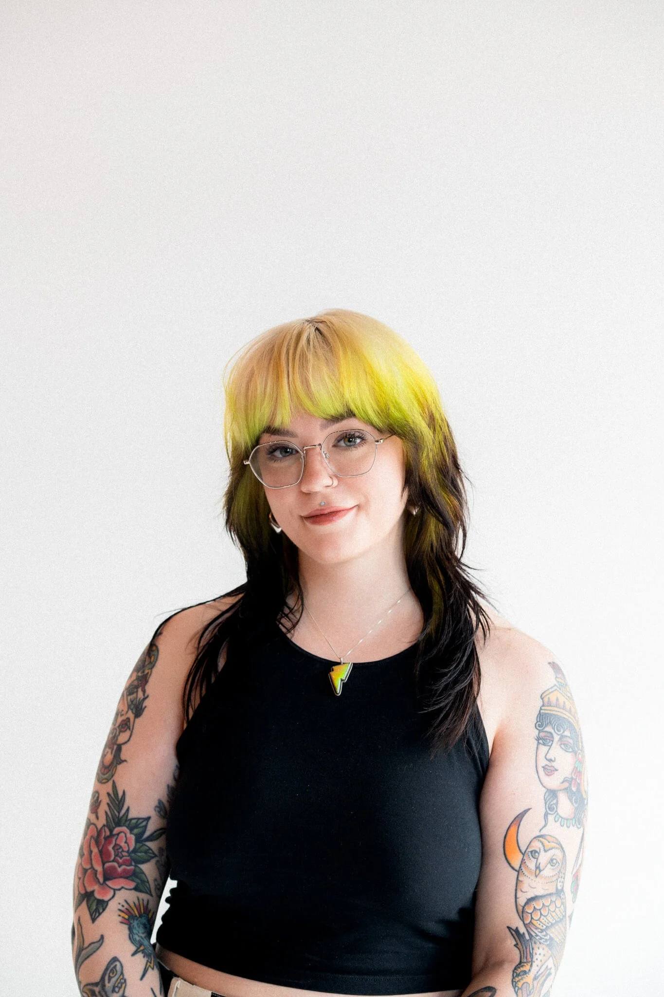 Marci, web and brand designer, is centered in the frame and has blonde, green, and black hair. They are wearing a black tank top and silver glasses, with a green lightning bolt necklace.