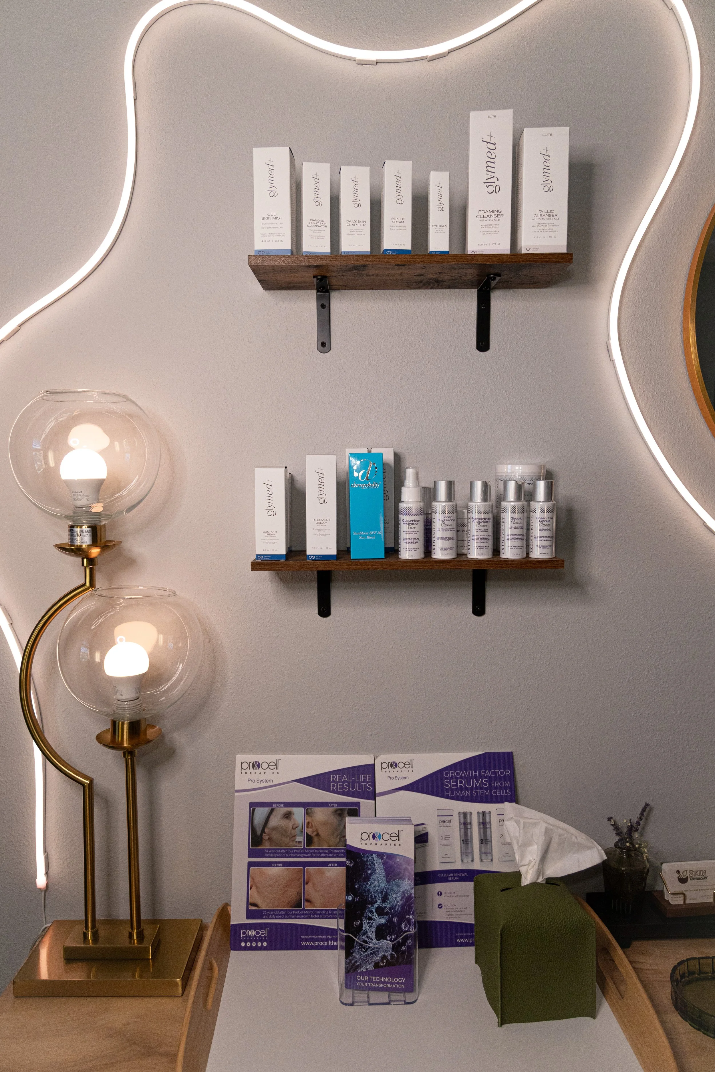 Bathroom or skincare clinic display with skincare products on wooden shelves, a lamp with glass globes, informational brochures on a desk, and a green chair.