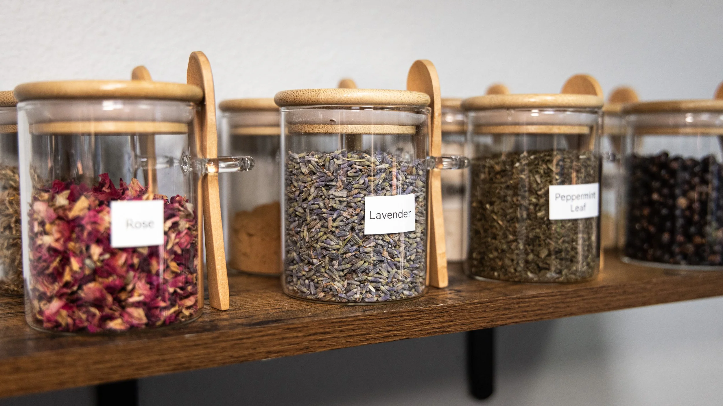 Shelf with glass jars containing dried herbs labeled Rose, Lavender, Peppermint Leaf, and other herbs.
