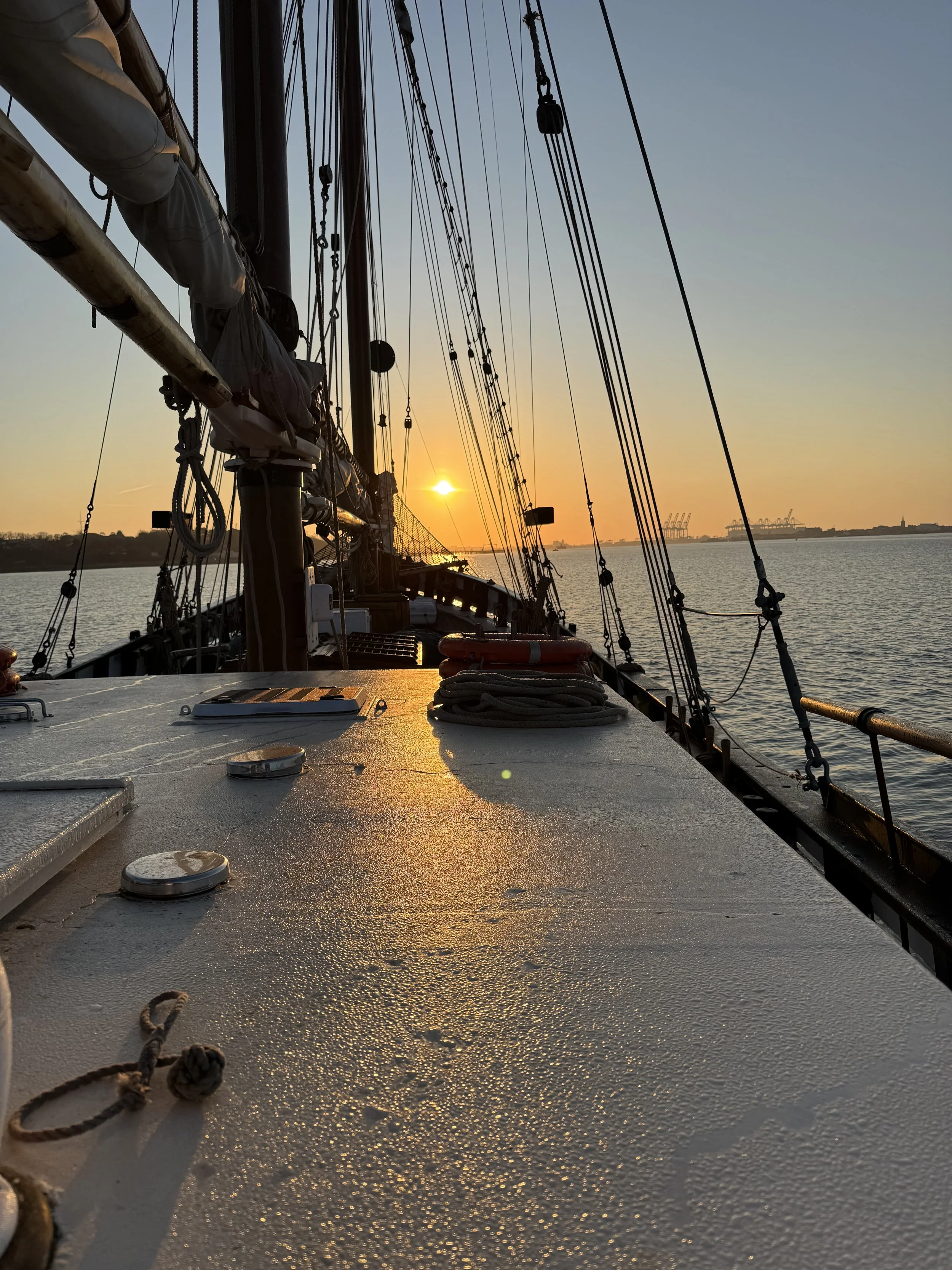 View from the deck of a sailboat at sunset, showing the rigging, ropes, and parts of the deck, with the water and distant shoreline visible.