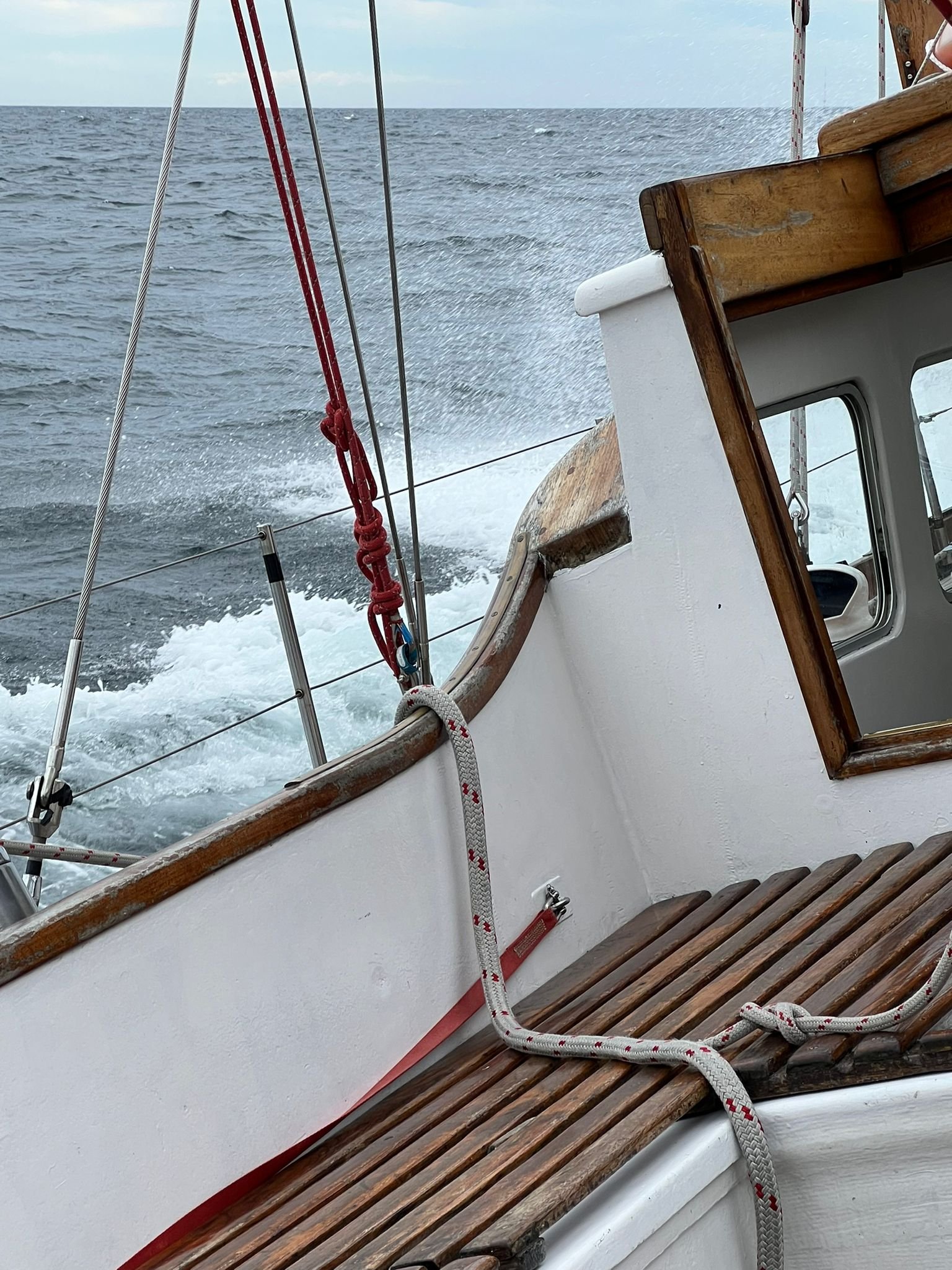 View from a sailboat showing water splashing behind as the boat moves, with ropes, a wooden railing, and a small window on the boat's cabin.