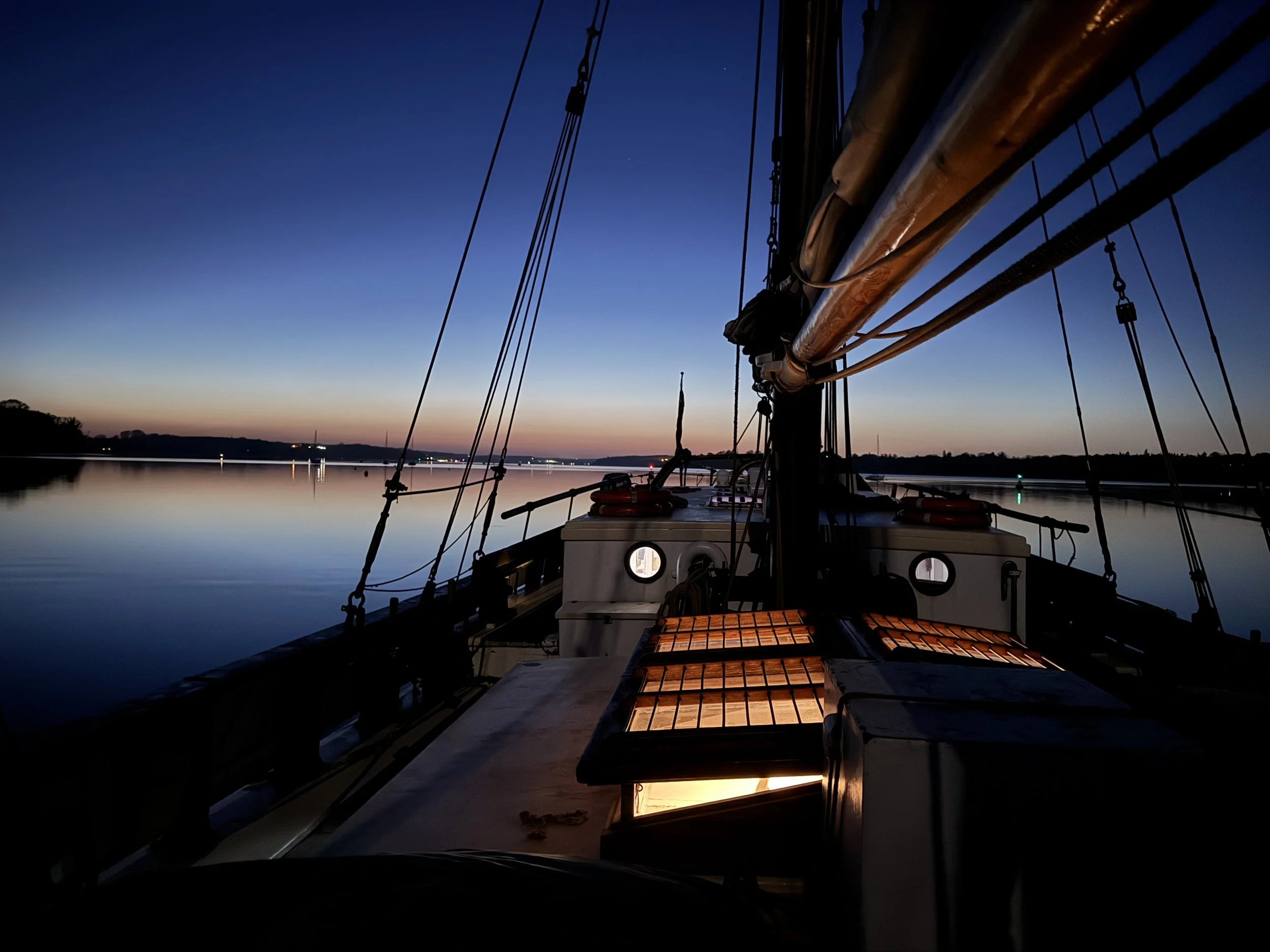 Looking down the deck of a sailboat at dusk, with a calm body of water and a colorful sunset sky in the background.