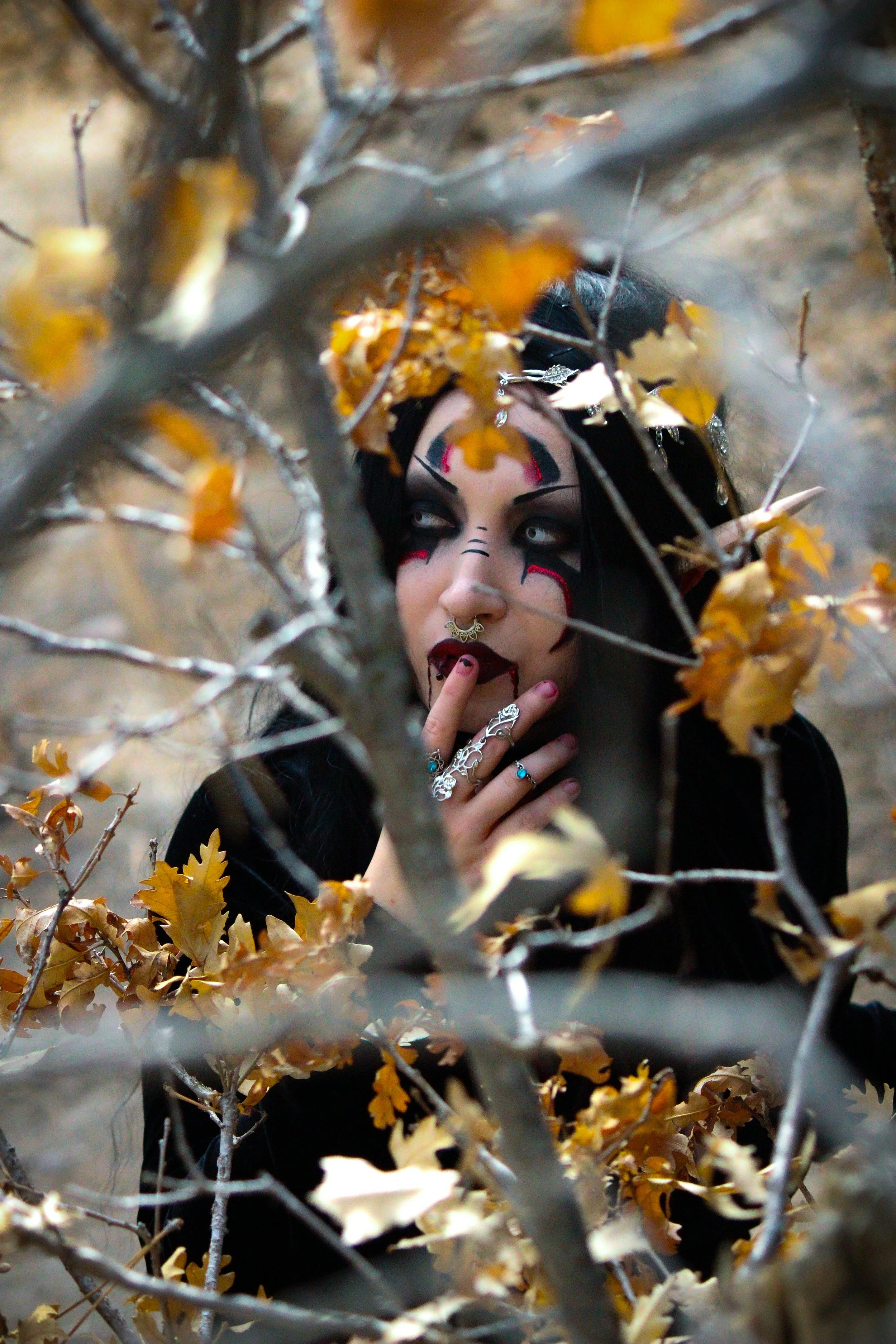 A person with gothic makeup and jewelry is behind a tree with dry leaves, peering through the branches and leaves.