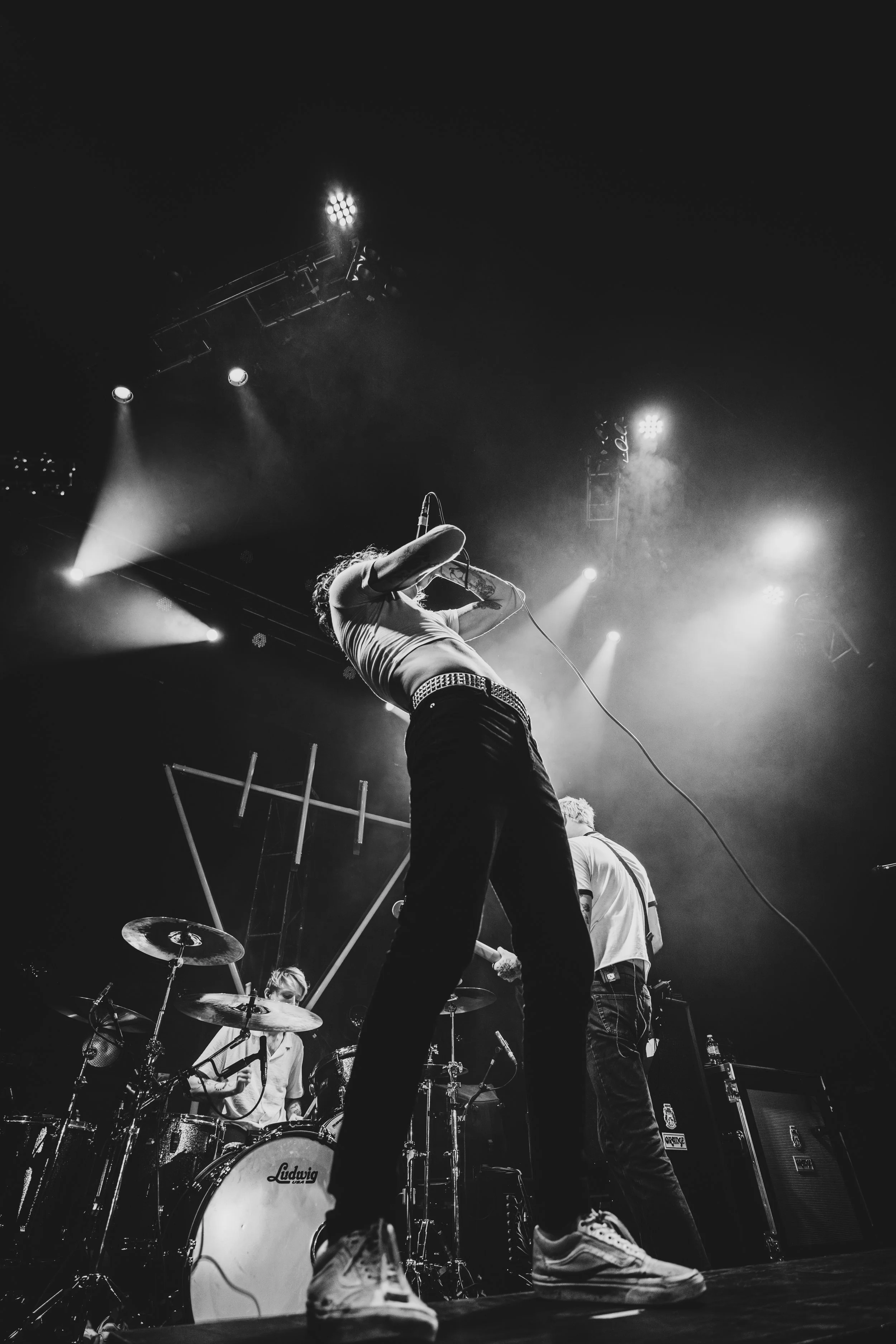 A black and white photo of a live band performance, featuring a vocalist singing into a microphone and a drummer playing drums on stage with stage lights overhead.