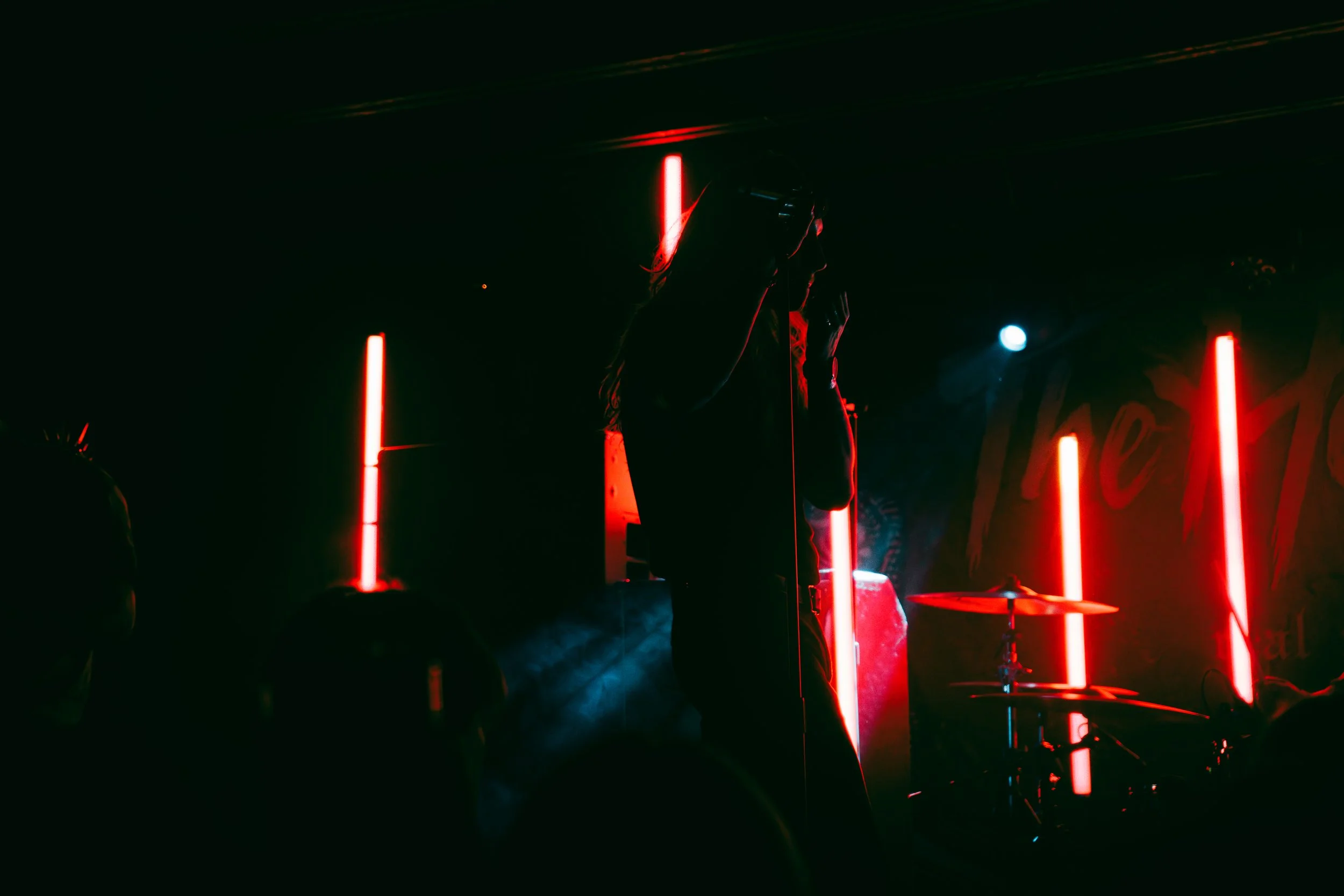 Silhouette of a singer on stage with red vertical neon lights and a drum set in the background, in a dark music venue.