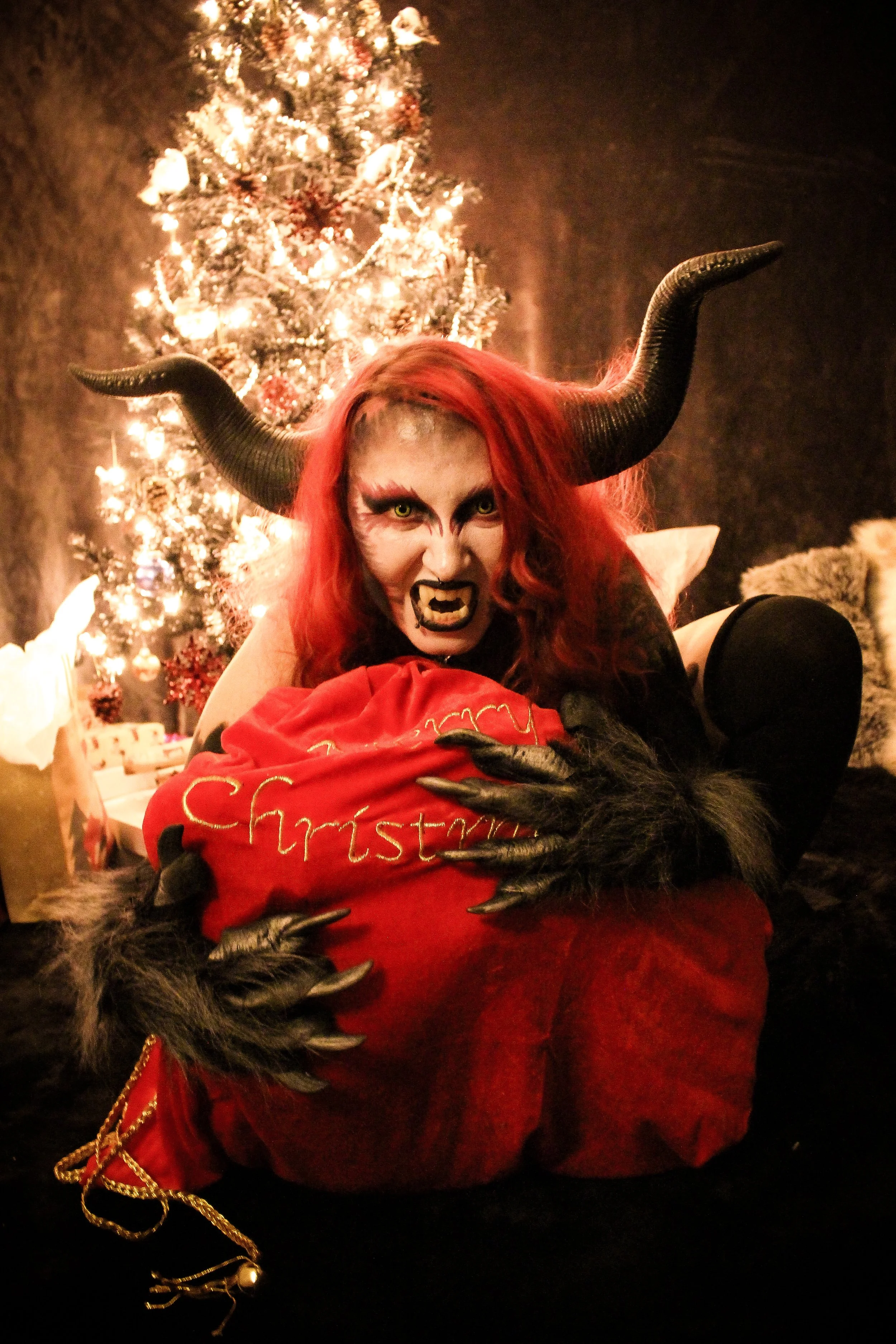 A person dressed as a devil with red hair, horns, and a spooky face, hugging a large red Christmas stocking in front of a decorated Christmas tree.