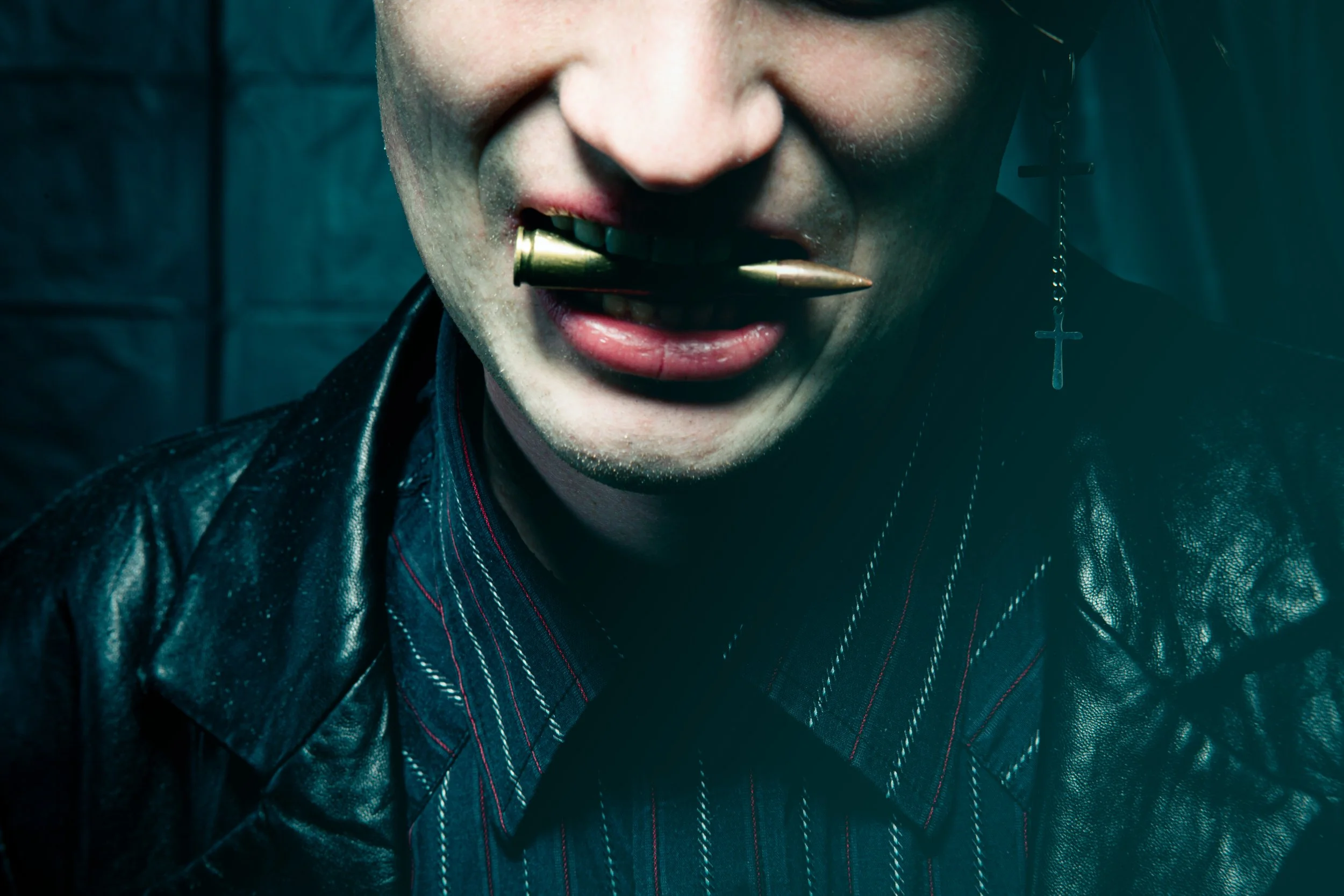 Close-up of a person's mouth with a bullet between their teeth and a gun in their mouth, wearing a leather jacket and a cross earring.