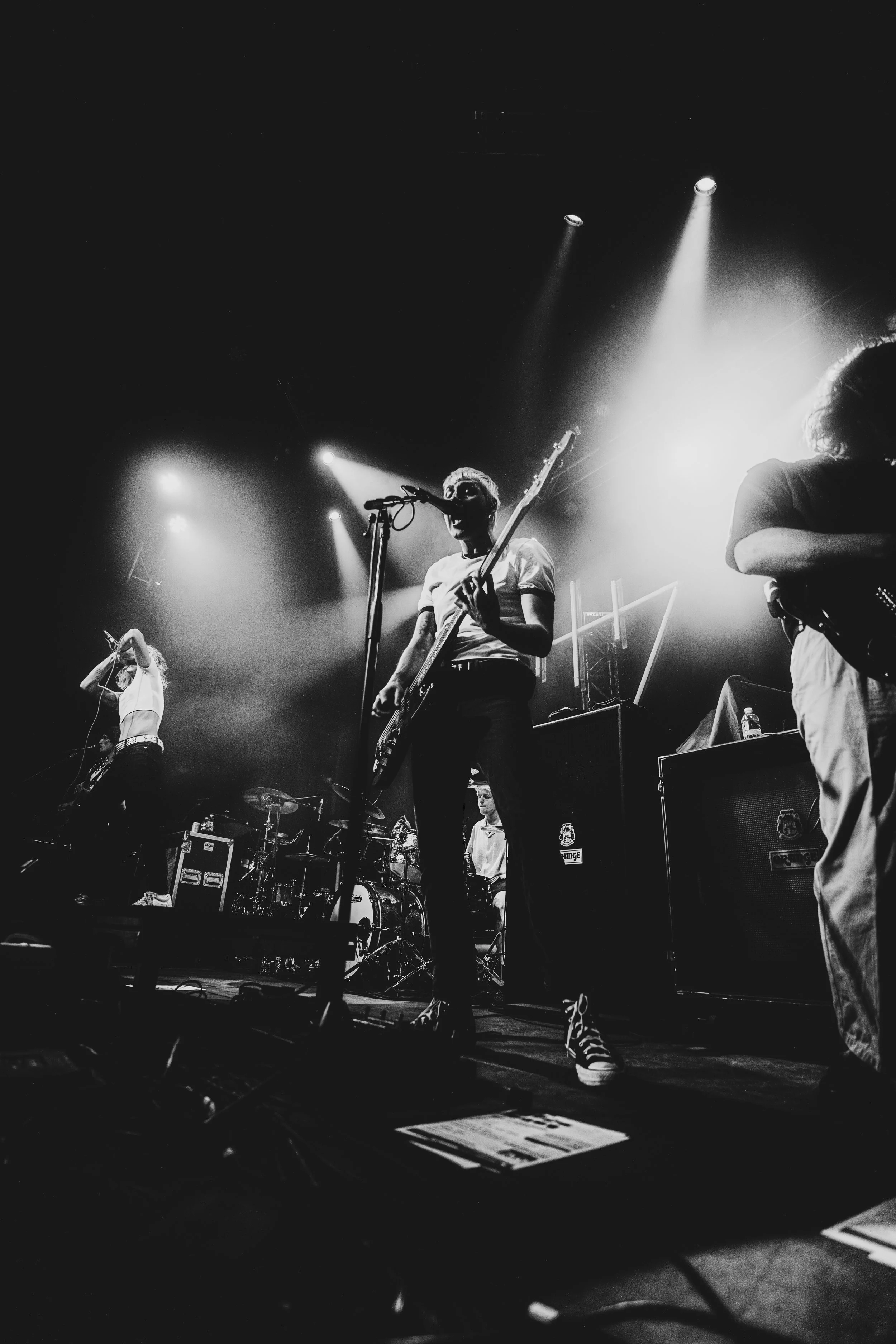 Black and white photo of a band performing on stage with bright spotlights overhead. The foreground features a male guitarist singing into a microphone, wearing a white t-shirt and striped shoes. To his left, another band member is seen singing with 