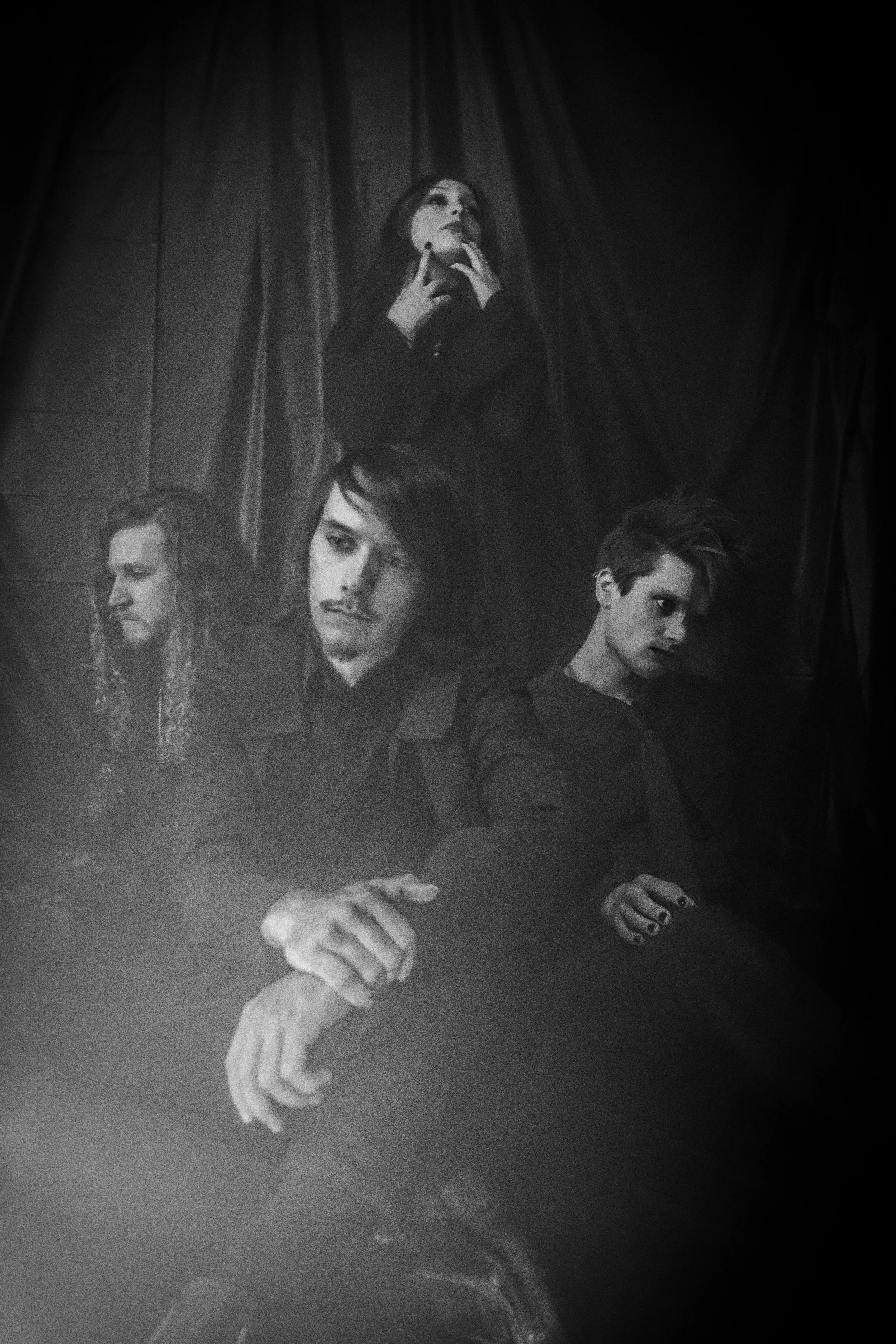 A black and white photo of four people posing against a dark curtain backdrop. The woman at the top is standing with her hands near her face, while the three people at the bottom are sitting, with one resting his arm over another's, all with serious 