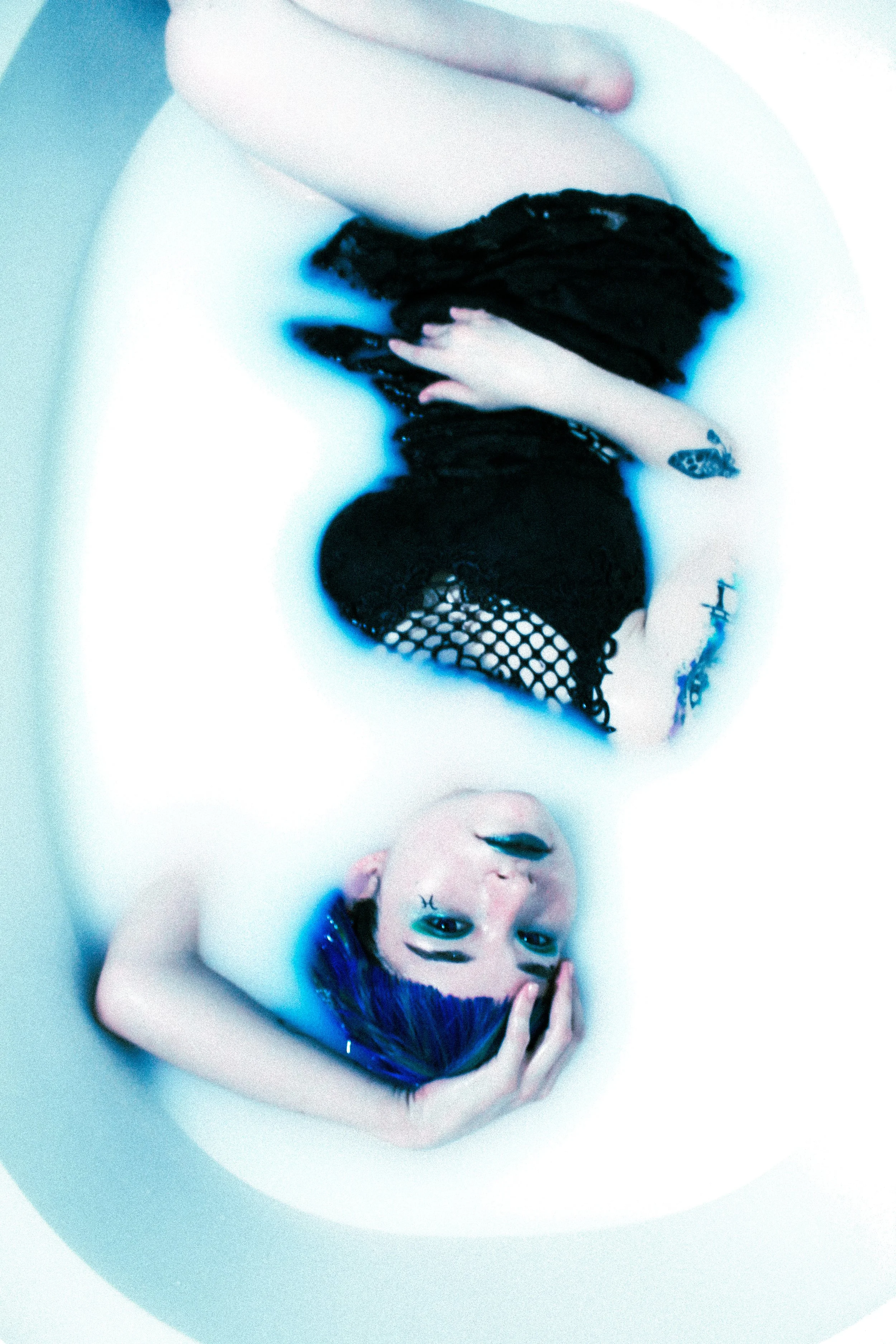 A woman with purple hair lying in a bathtub filled with milky water, wearing a black fishnet garment.
