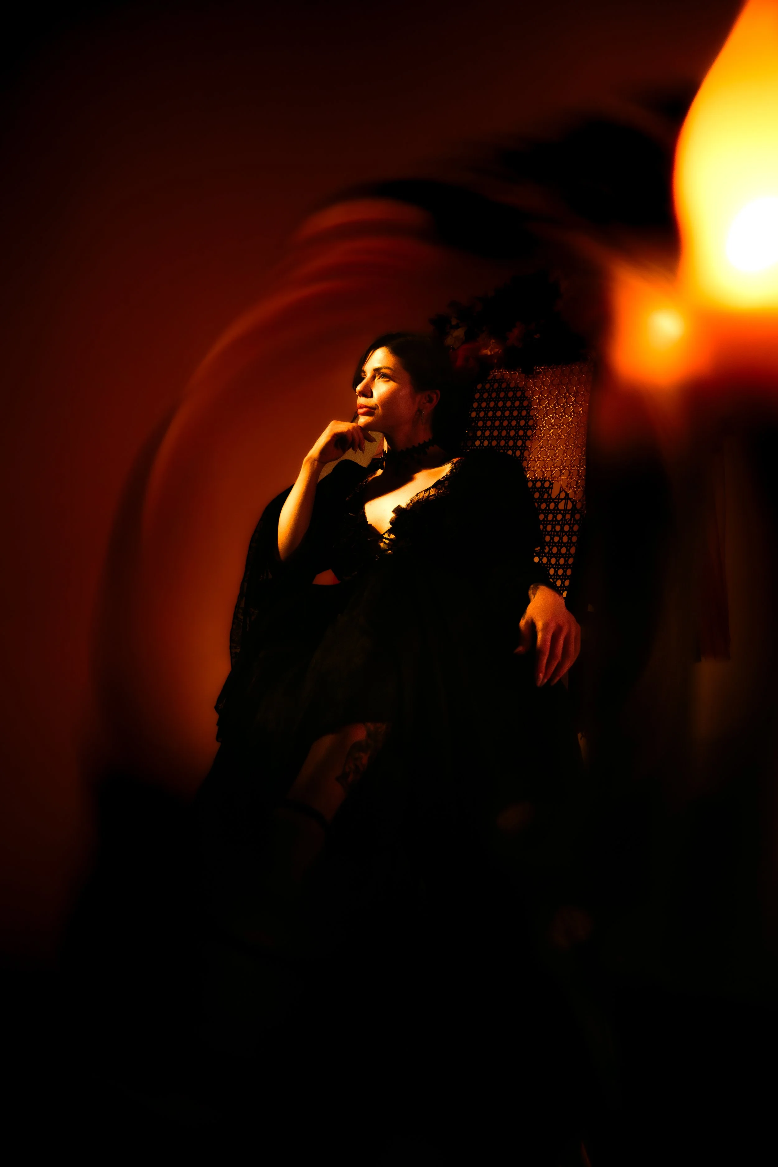 A woman wearing a black lace dress, sitting on a chair in a dimly lit room with warm lighting.