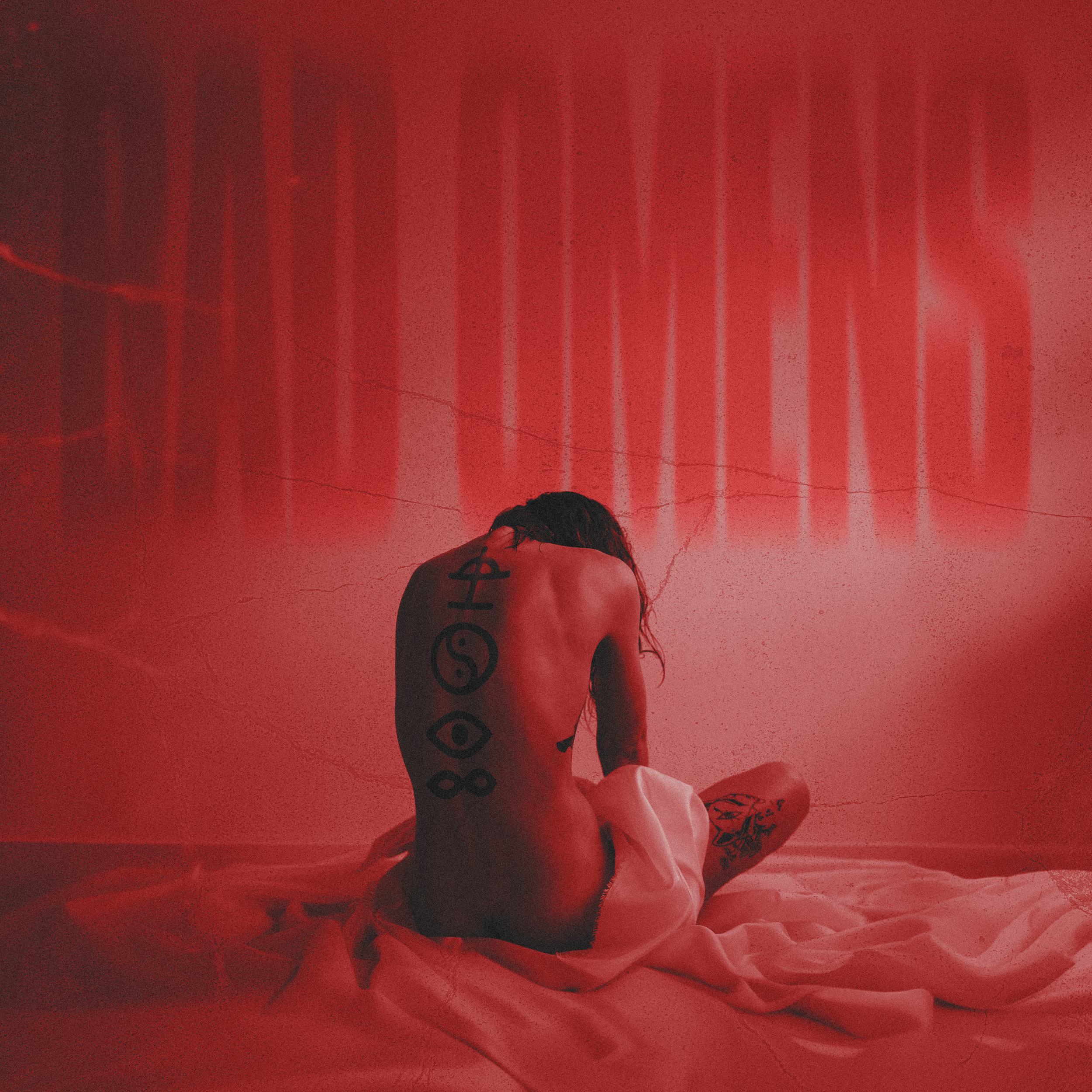 A person with tattoos sitting on a bed with their back facing the camera, illuminated by red lighting and a textured wall in the background.