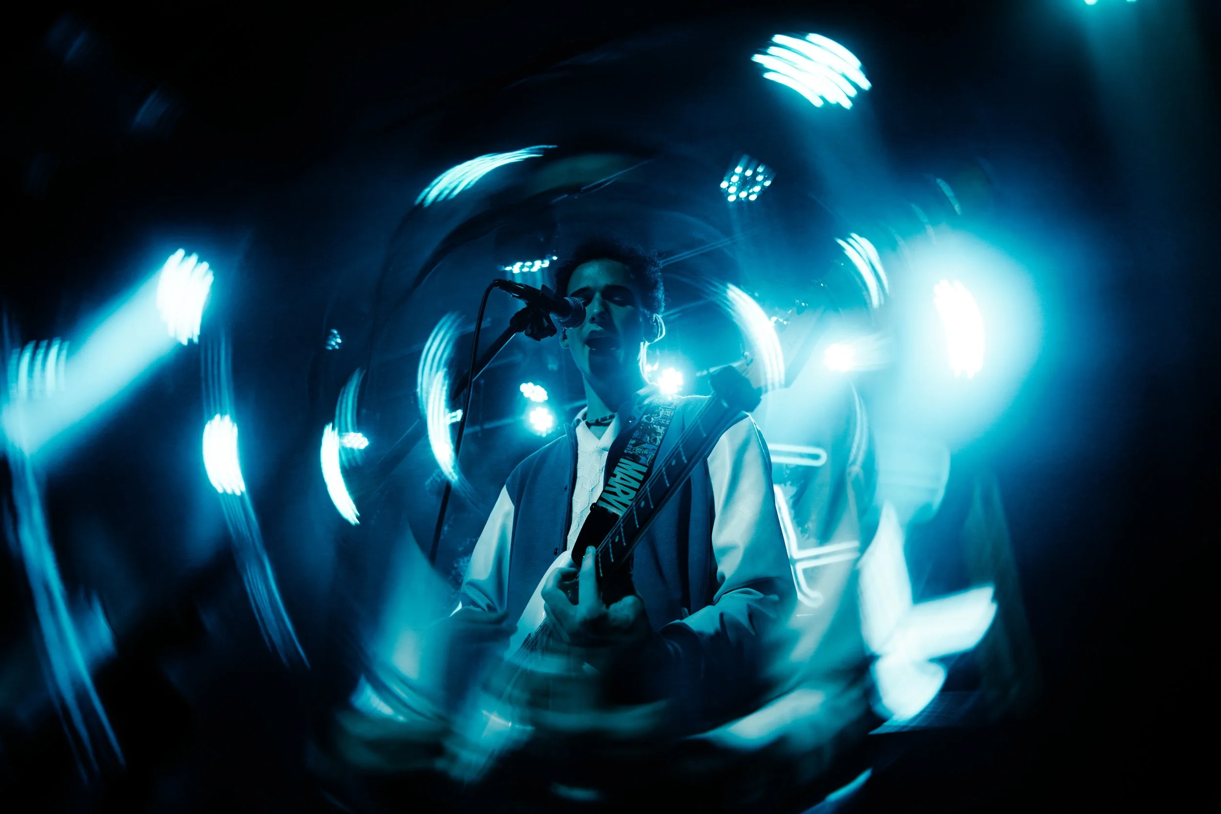 A musician performs on stage with blue lighting, singing into a microphone and playing an electric guitar, surrounded by abstract light streaks.