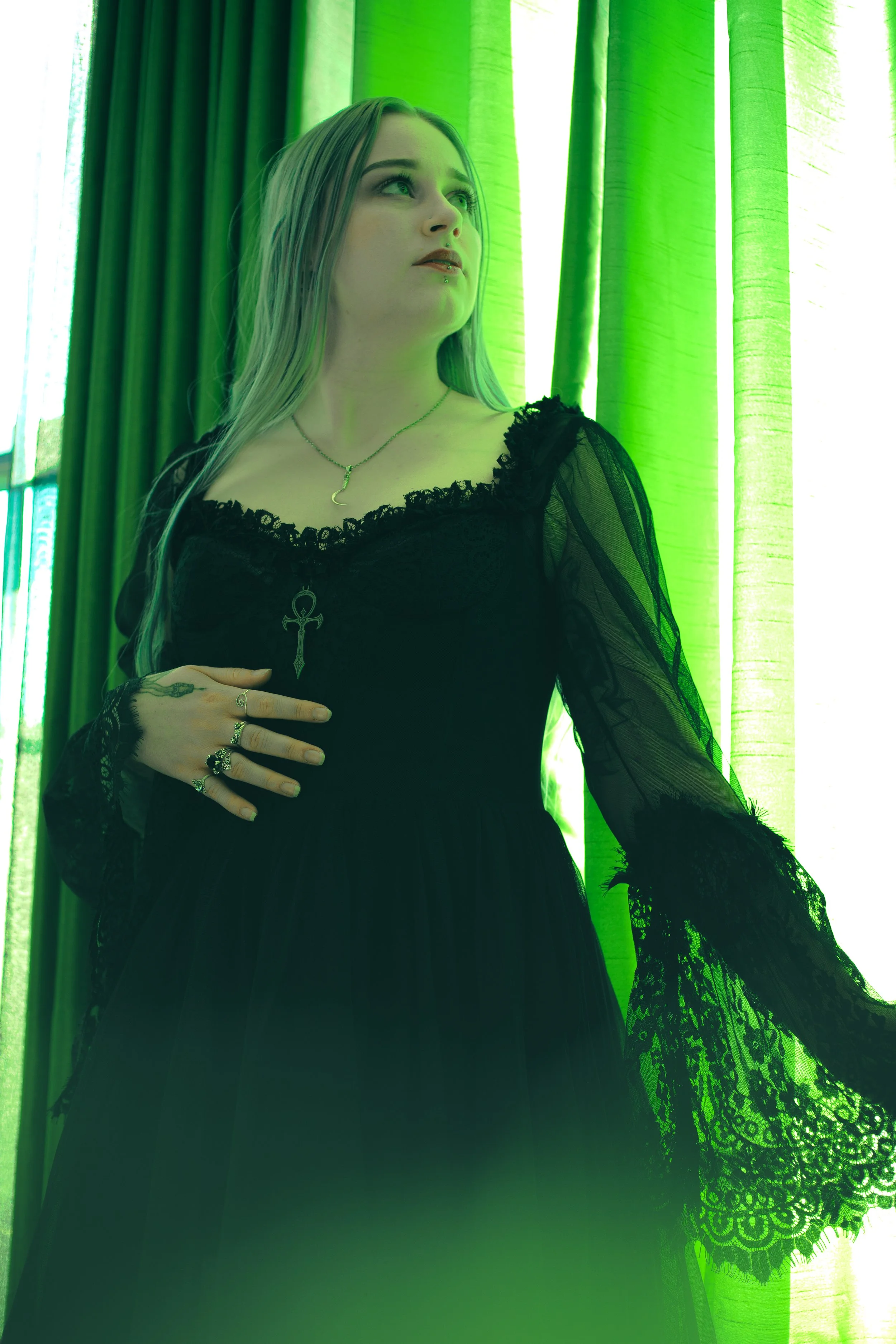 A woman with long blonde hair wearing a black lace dress with sheer sleeves, standing near green curtains, looking to her right.