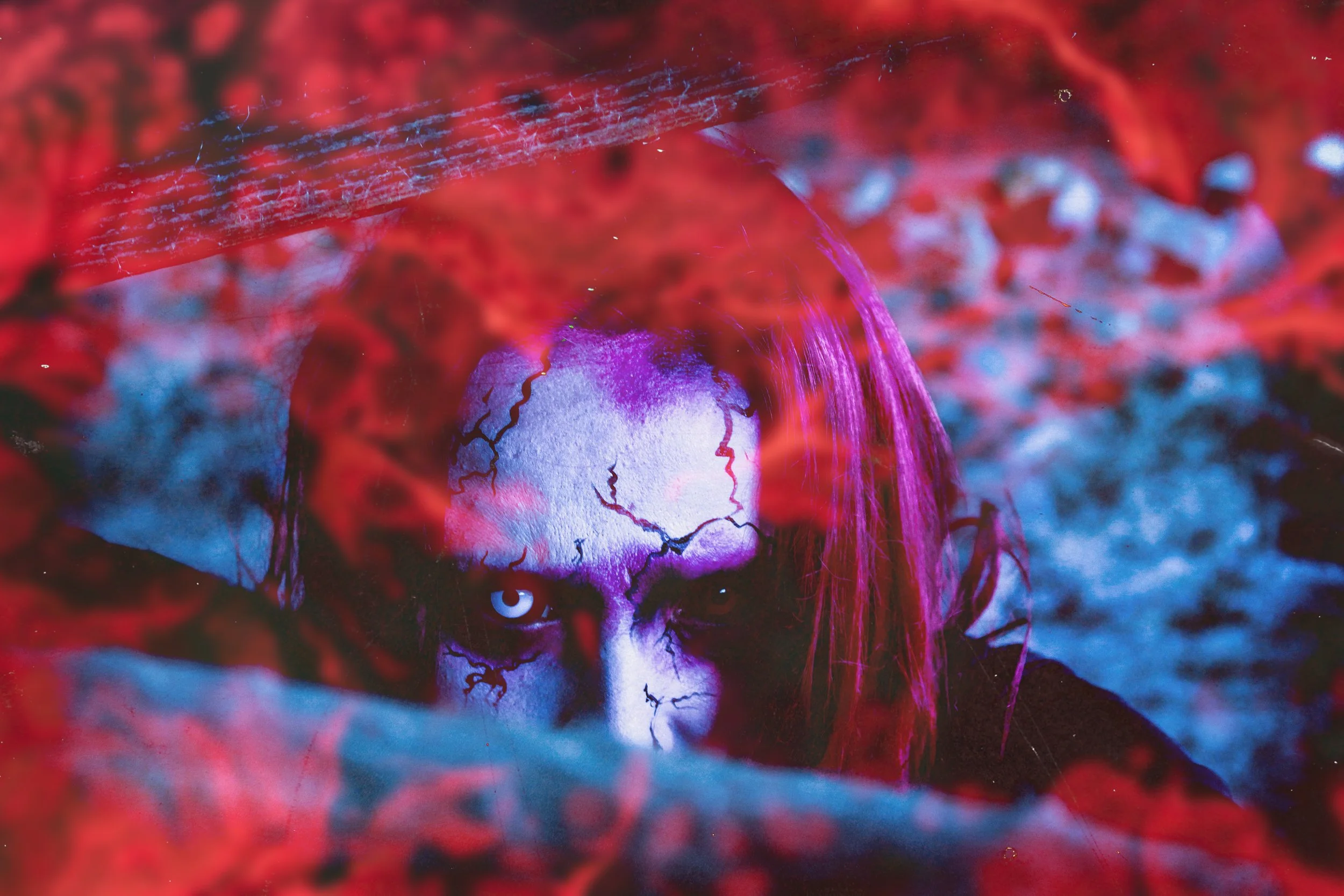 Person with white face and cracked face paint, intense gaze, surrounded by red and purple elements, possibly part of a themed event or costume.