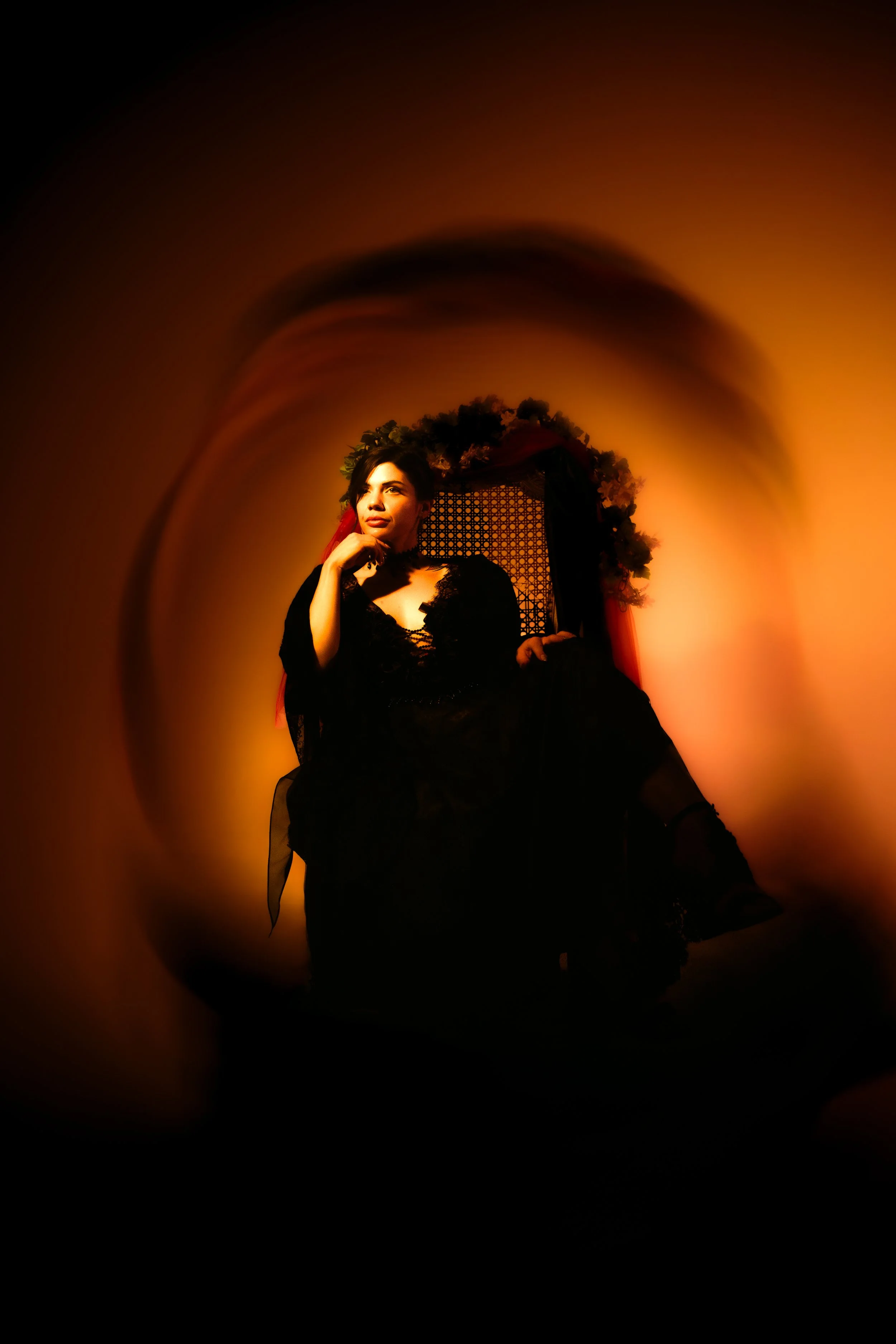 A woman in a black dress sitting on a throne with a floral crown, illuminated by warm orange light.