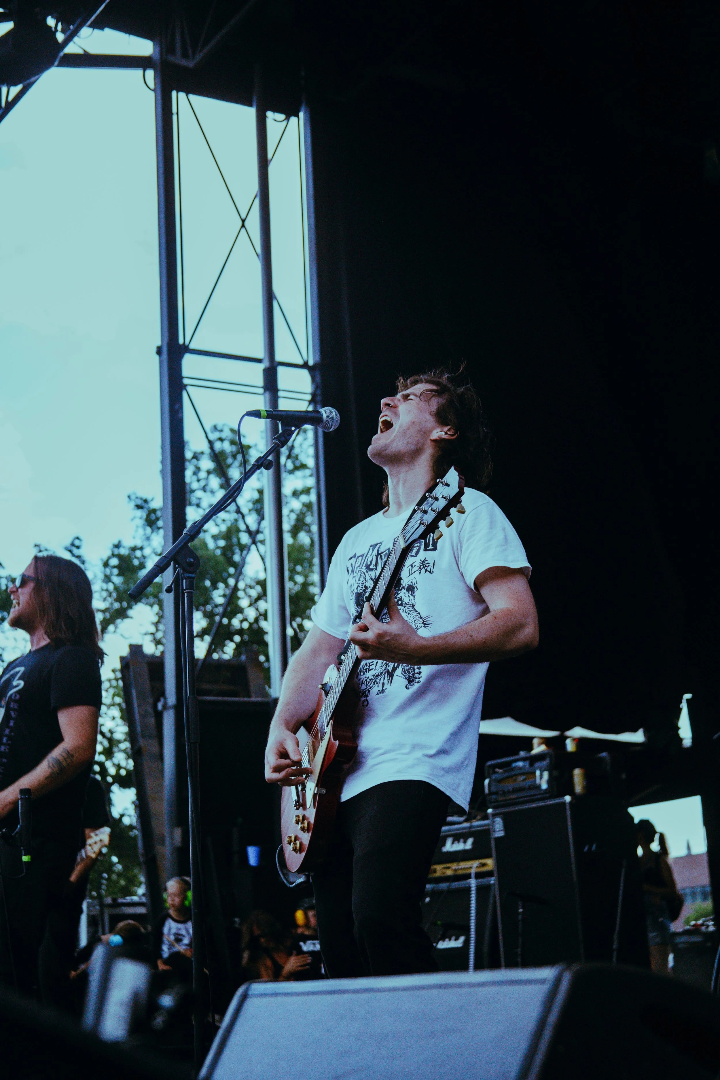 A male musician passionately singing and playing an electric guitar on stage at an outdoor concert, with a female musician nearby and a mixed crowd in the background.