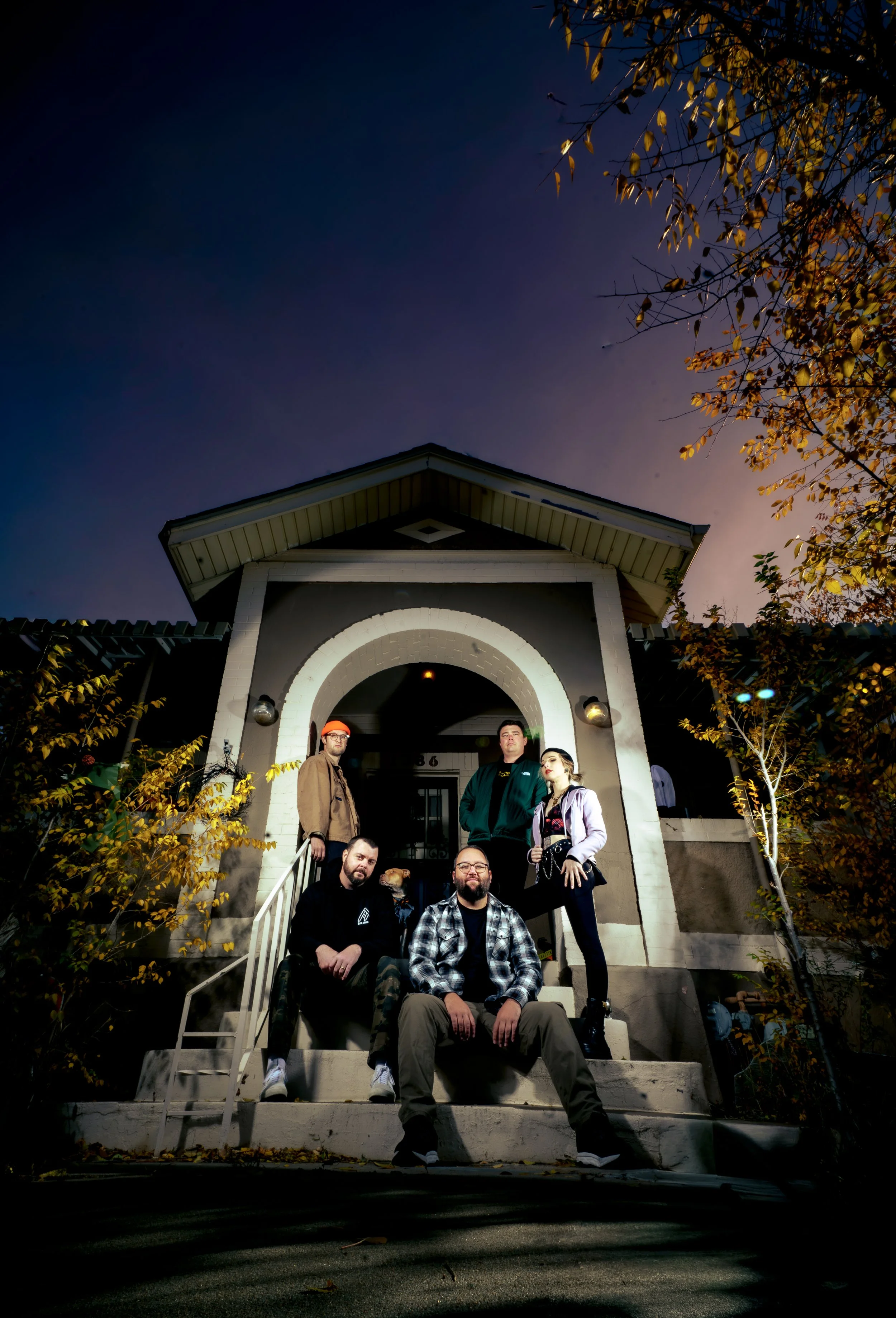 A group of six people sitting and standing on the steps of a house at night, with a dark sky and some autumn leaves on the trees.
