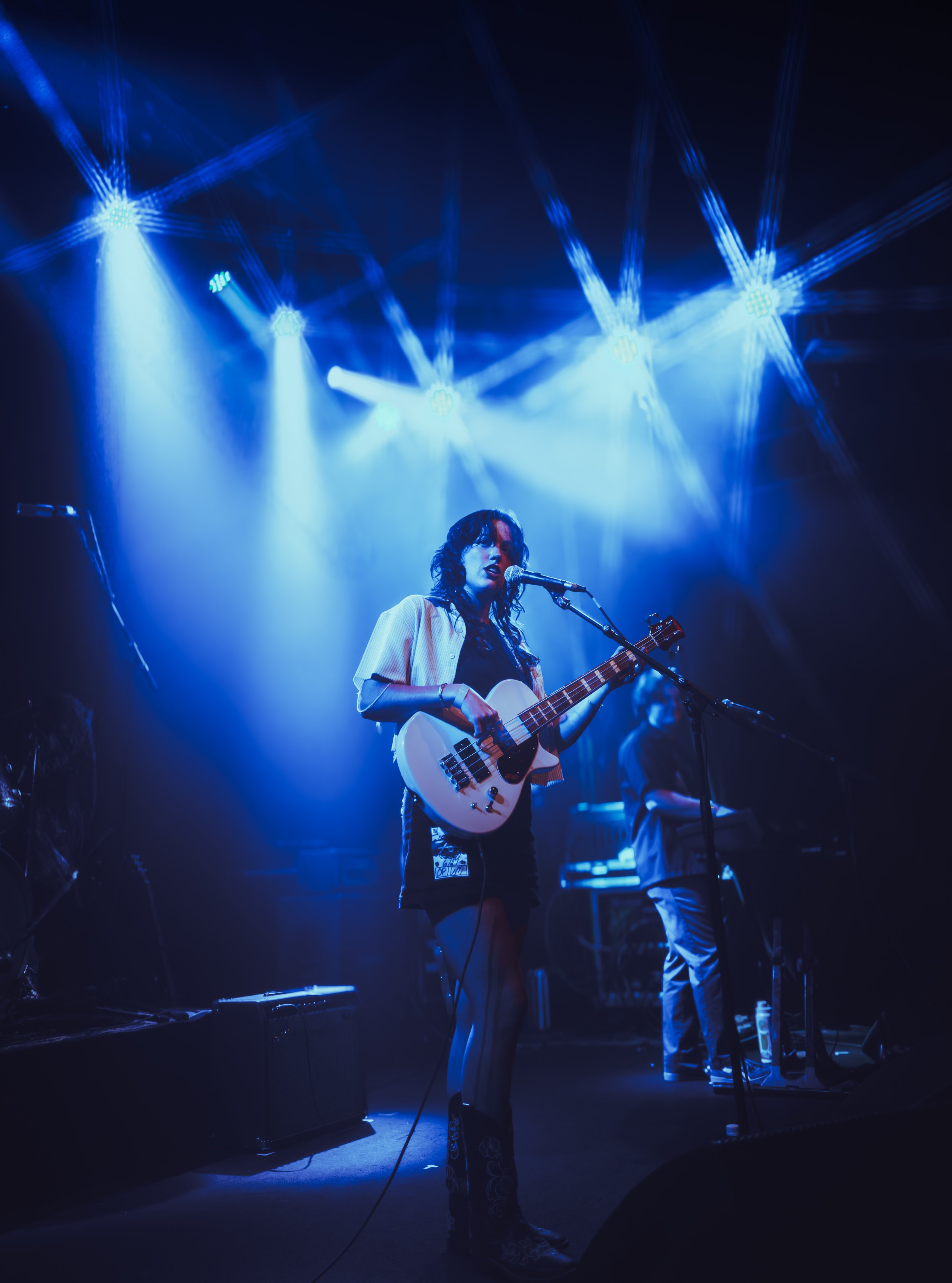A female musician performing on stage with a guitar, illuminated by blue stage lights, with a keyboard player in the background.