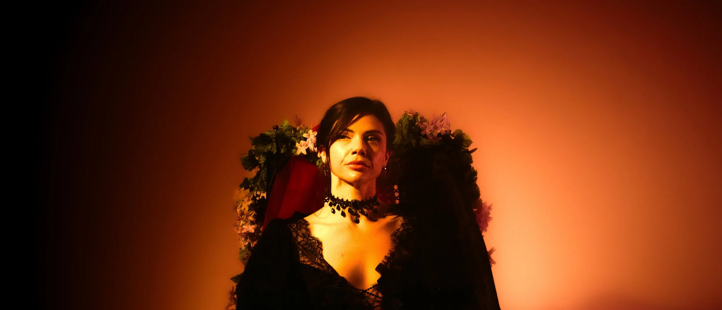A woman with dark hair wearing black lace clothing and a black statement necklace standing in front of a colorful floral background with warm orange and red lighting.