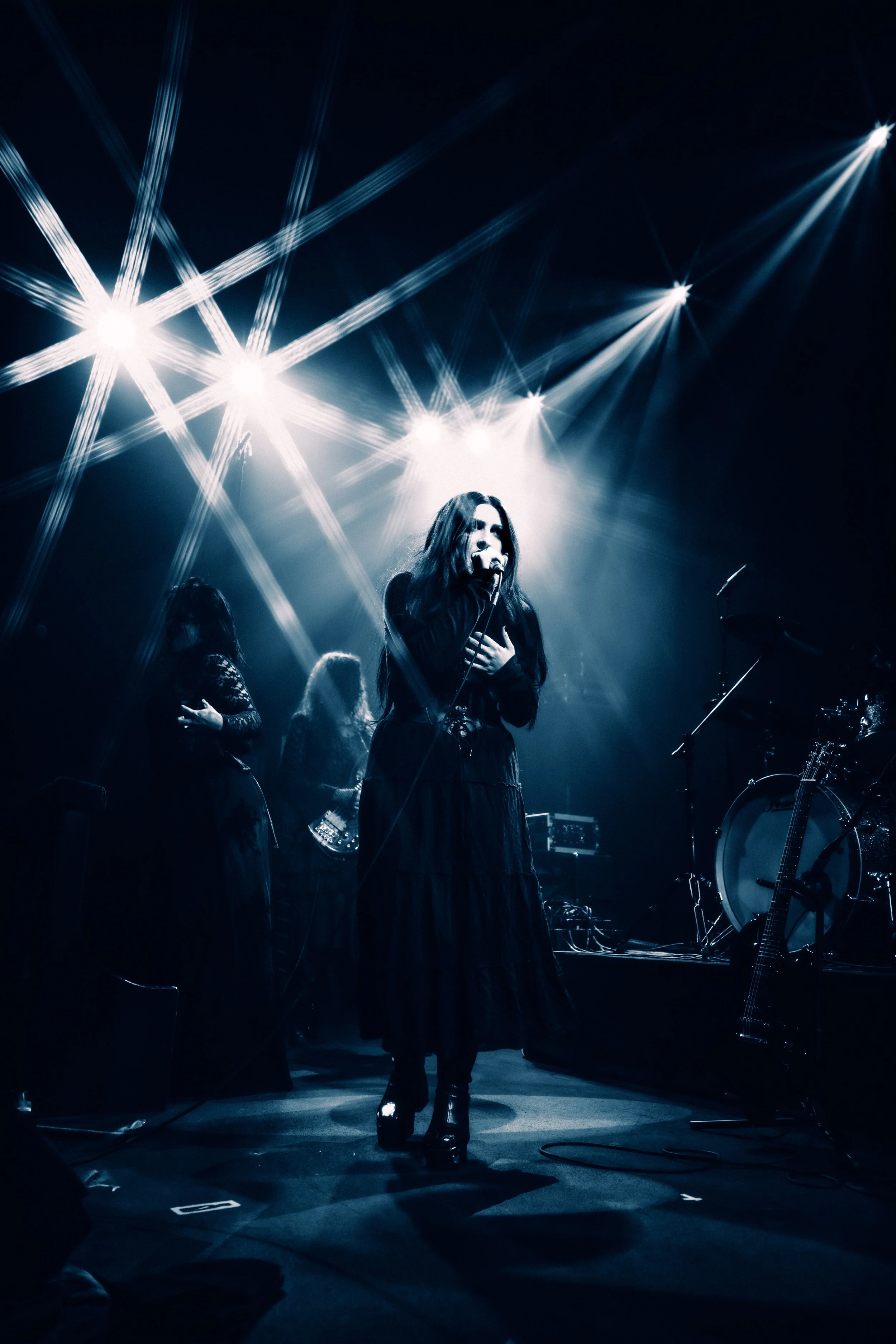 A female singer performing on stage with styling lighting effects, accompanied by two backup singers and musical instruments including a drum set and guitar.