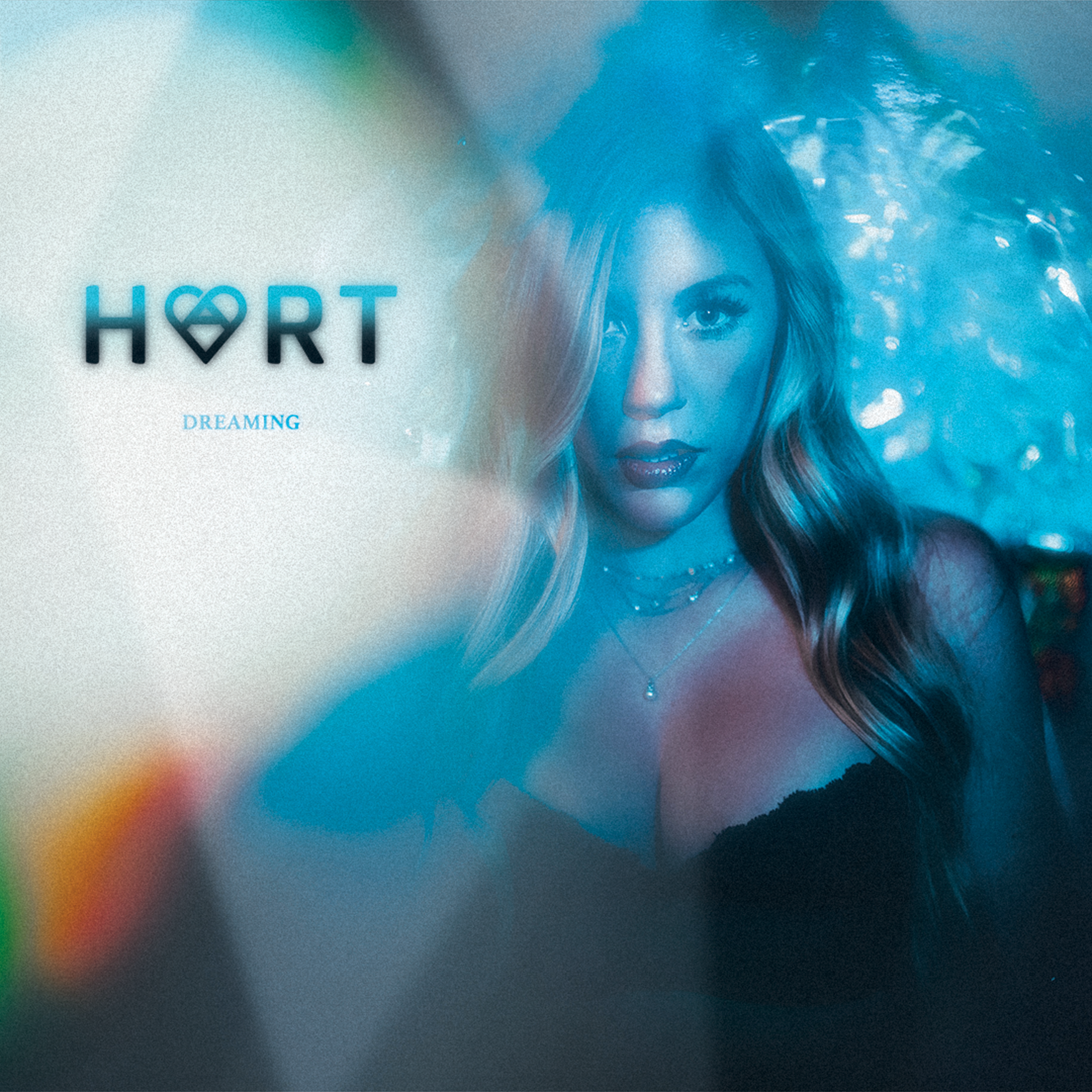A woman with long blonde hair and blue eyes, wearing dark lipstick and layered necklaces, is depicted in a blue-tinted, dreamy setting. The left side of the image shows the word "HØRT" with a heart-shaped design in place of the second "O" and the wor