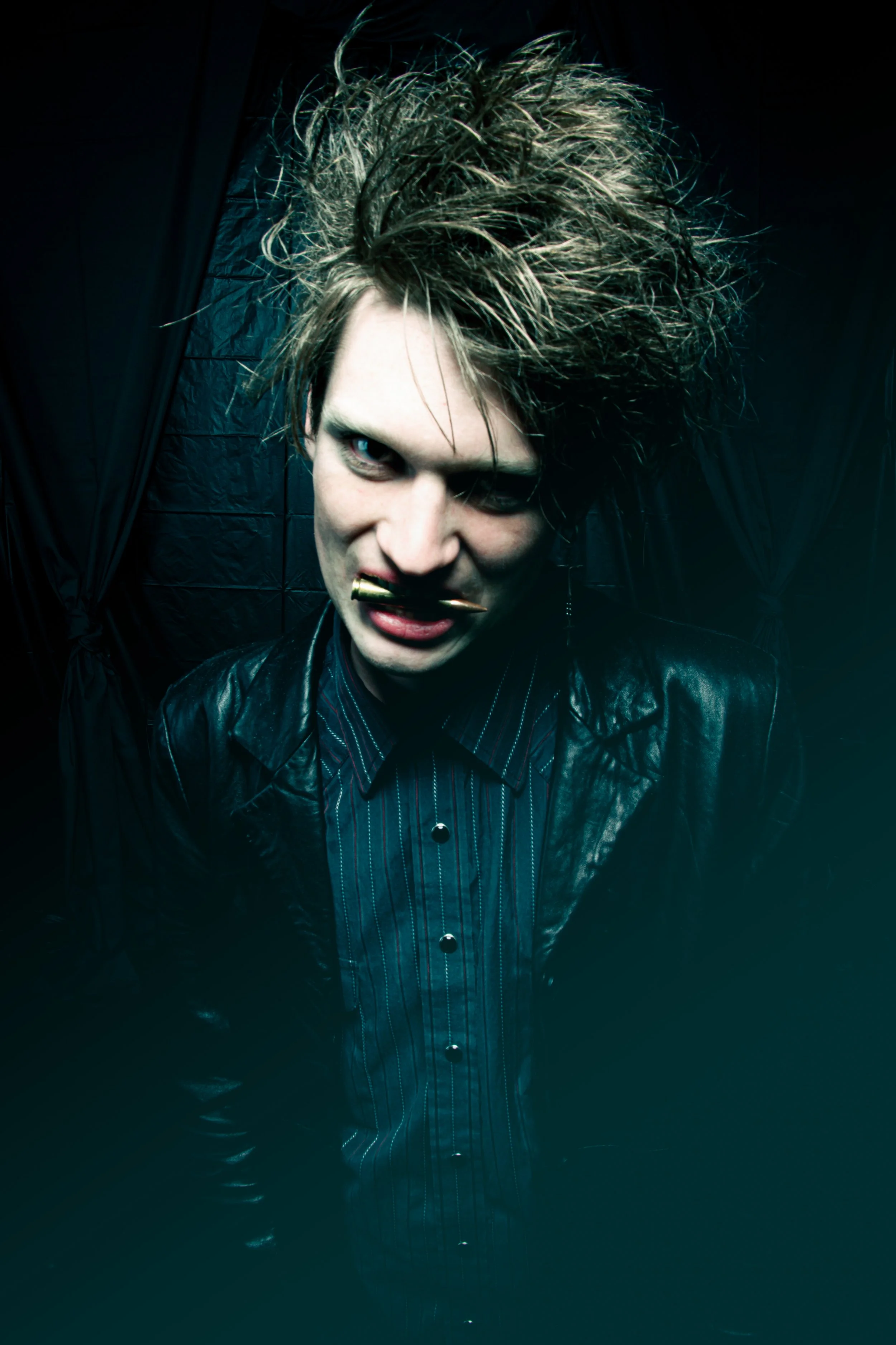 A person with spiked hair, heavy eye makeup, and a leather jacket holding a pen in their mouth, standing against a dark background.