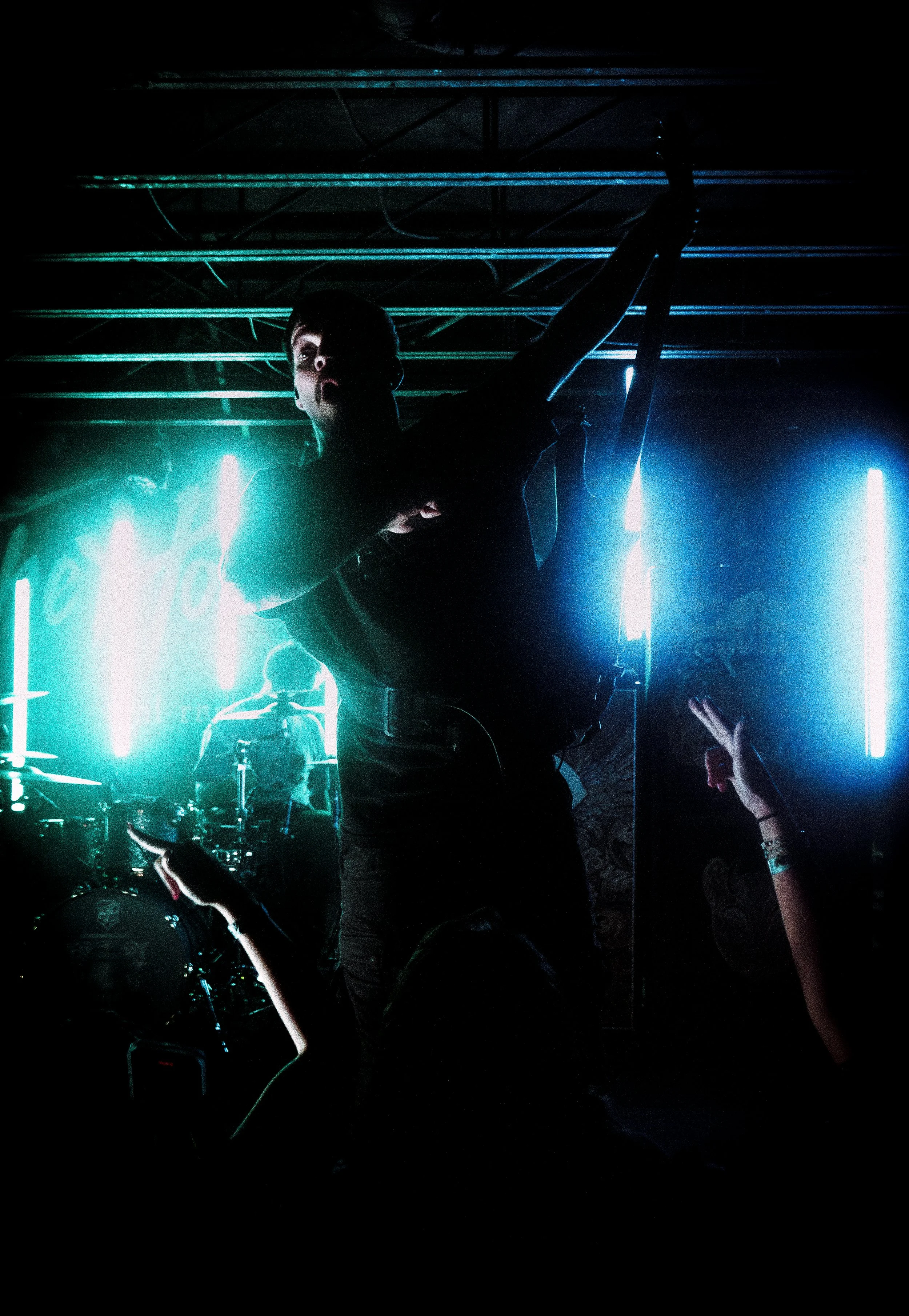 A musician performs on stage with a microphone, illuminated by blue and neon lights, with hands reaching up from the audience.