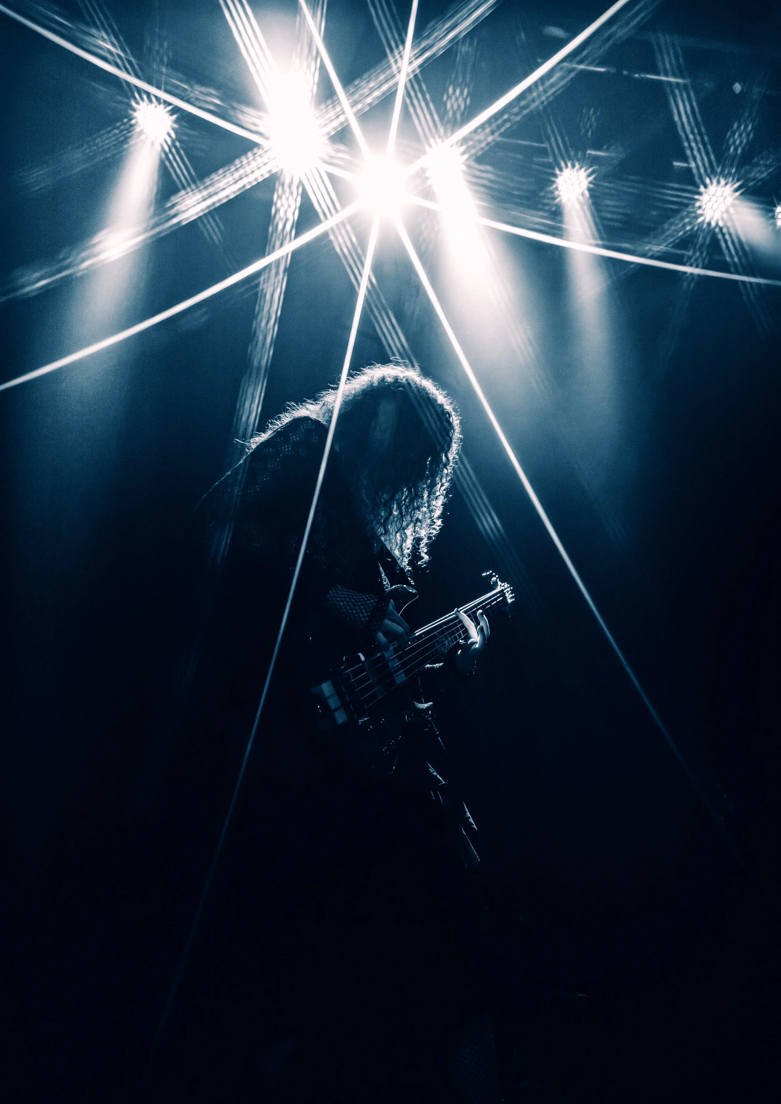 A female musician playing an electric guitar on stage, illuminated by bright stage lights creating starburst effects, with a shadowed background.