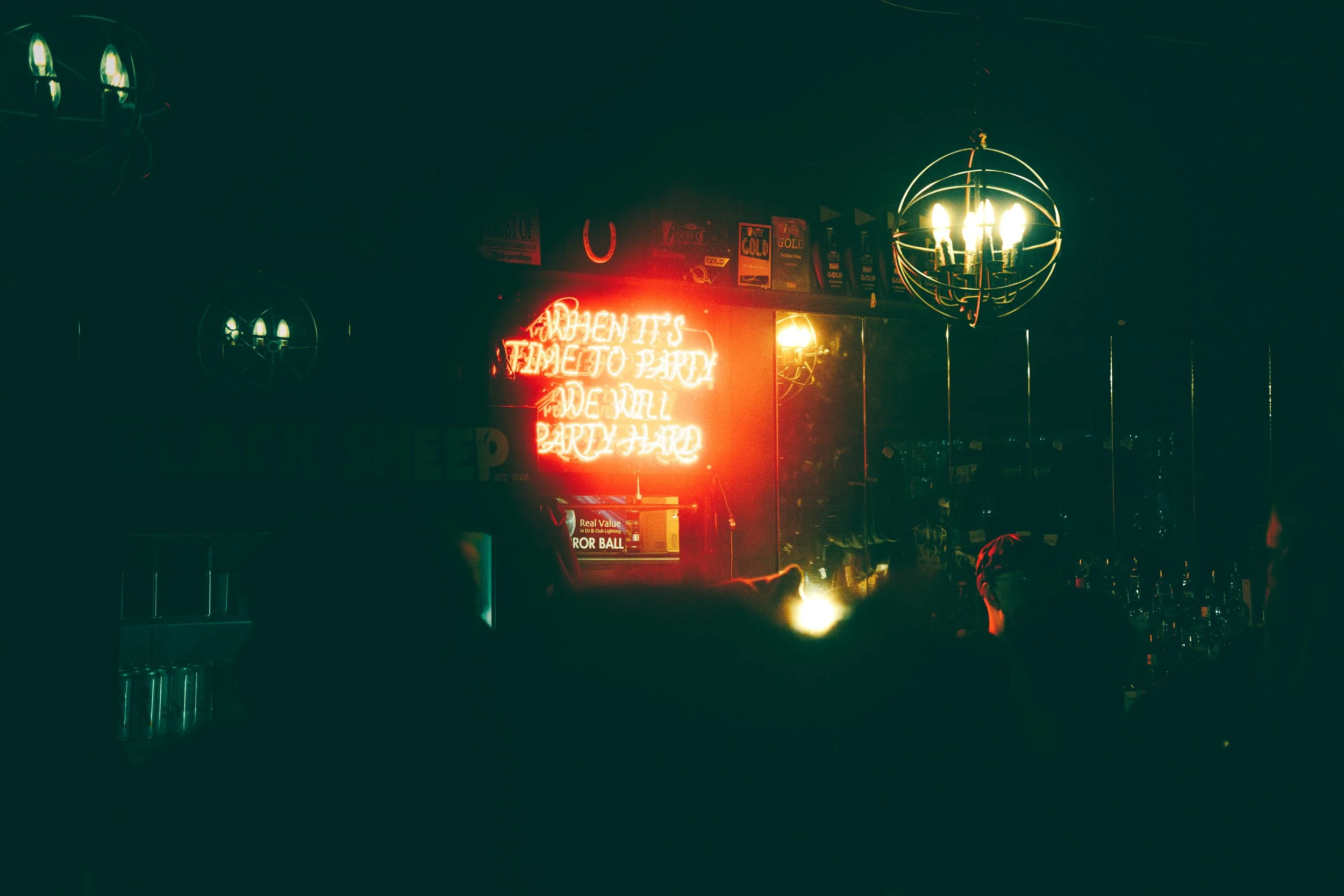 A dimly lit bar with a neon sign that reads 'When it's time to party, we will party hard,' with silhouettes of people and decorative lights hanging from the ceiling.