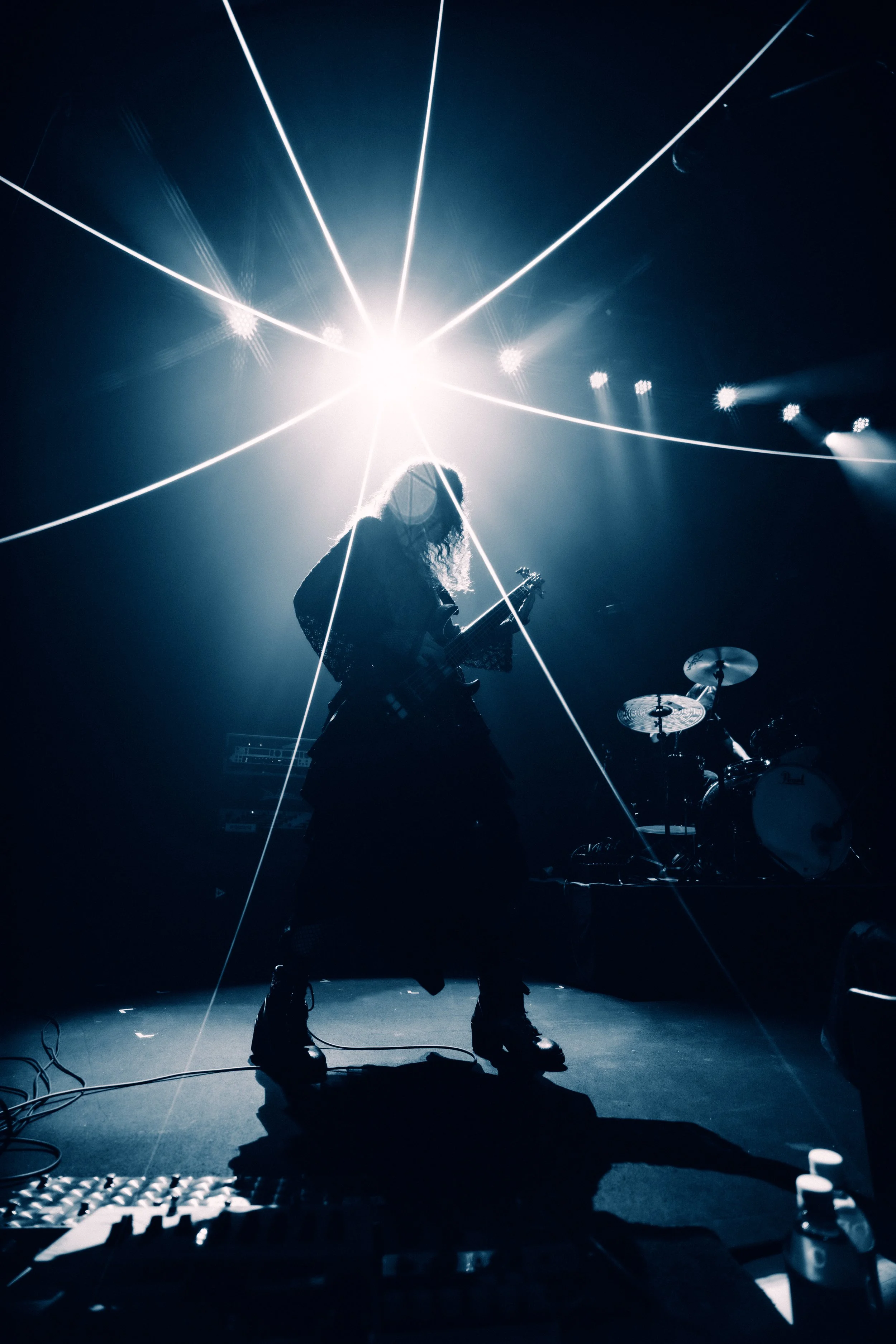 Silhouette of a female guitarist performing on stage with bright stage lights creating starburst effects around her, and a drummer visible in the background.