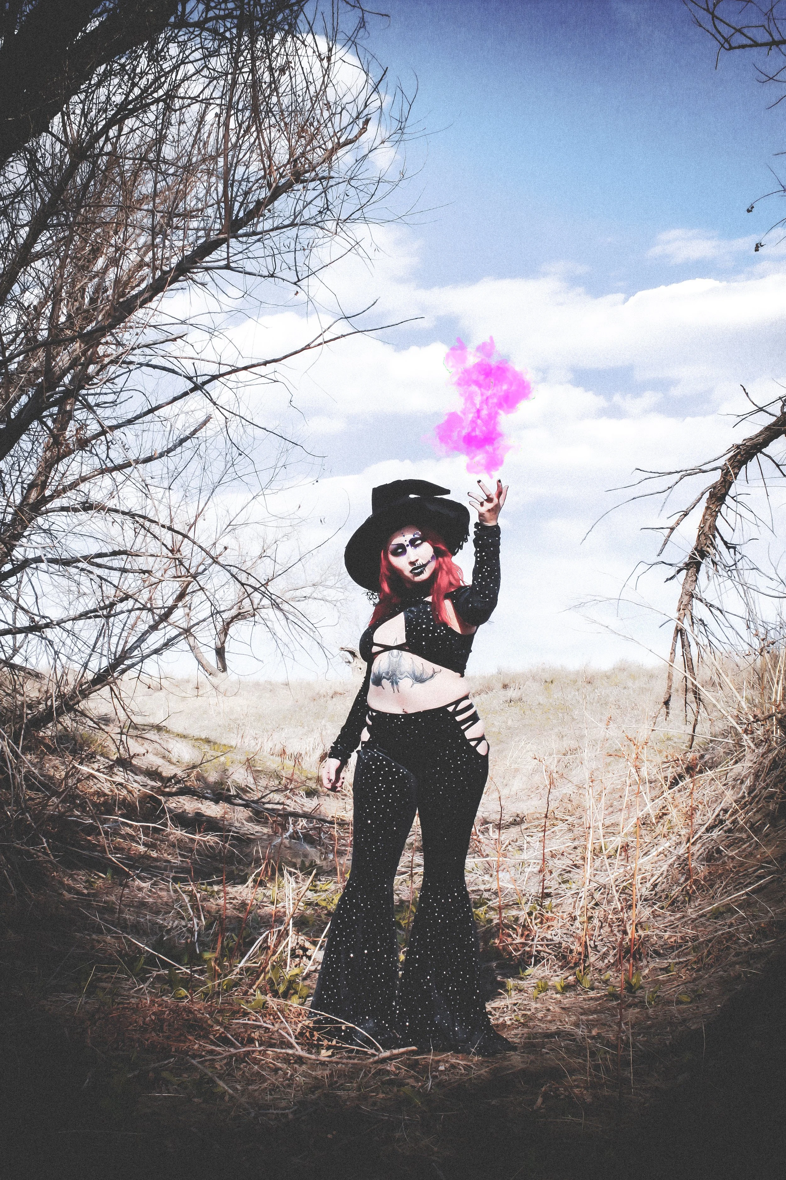 A woman with bright red hair dressed as a witch, wearing a black hat and black starry outfit, standing outdoors in a clearing with leafless trees and a bright cloudy sky, holding pink smoke in her hand.