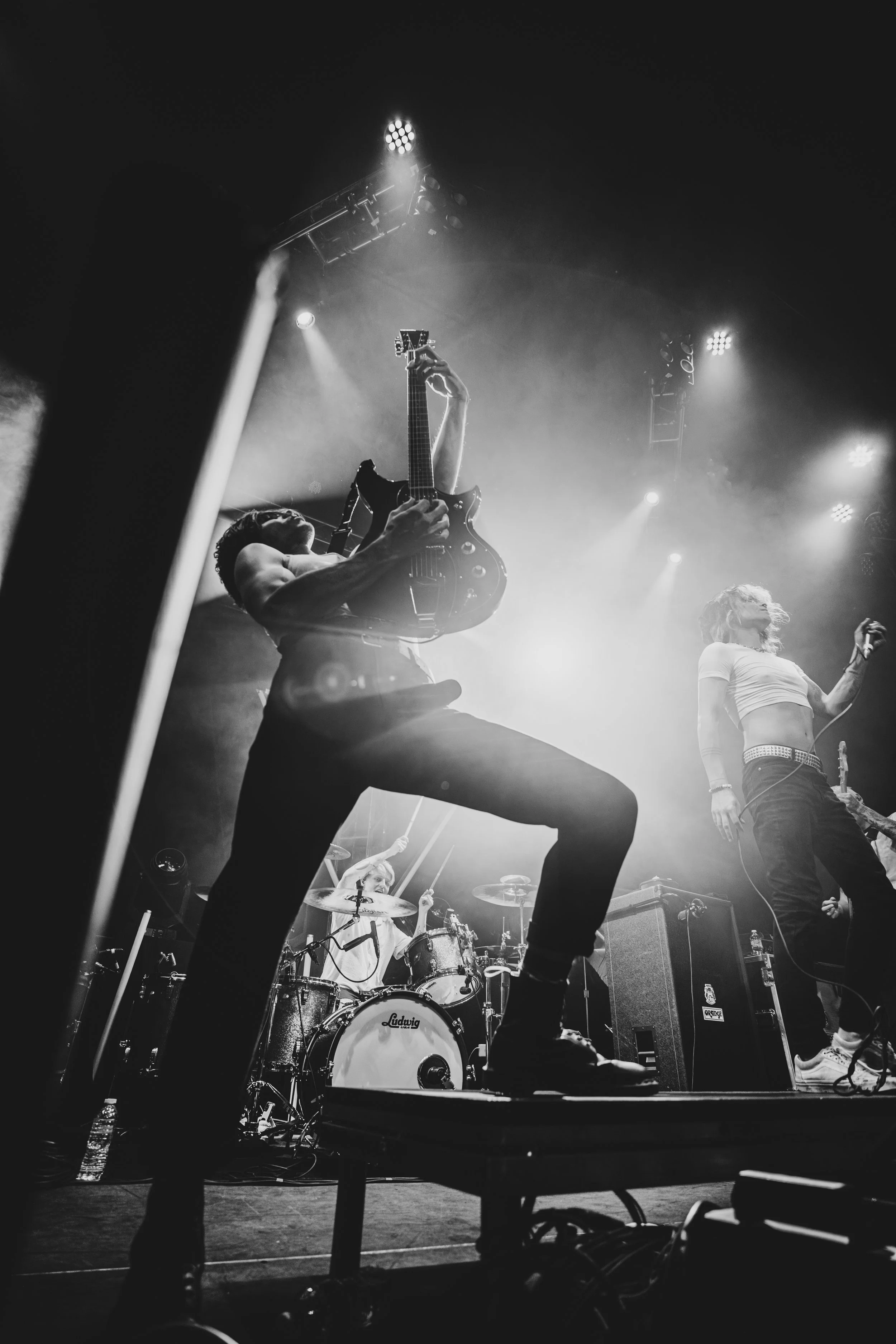 Black and white photo of a live rock band performance on stage, featuring a guitarist kneeling with one foot on a monitor and playing an electric guitar, a female vocalist holding a microphone, and a drummer in the background.