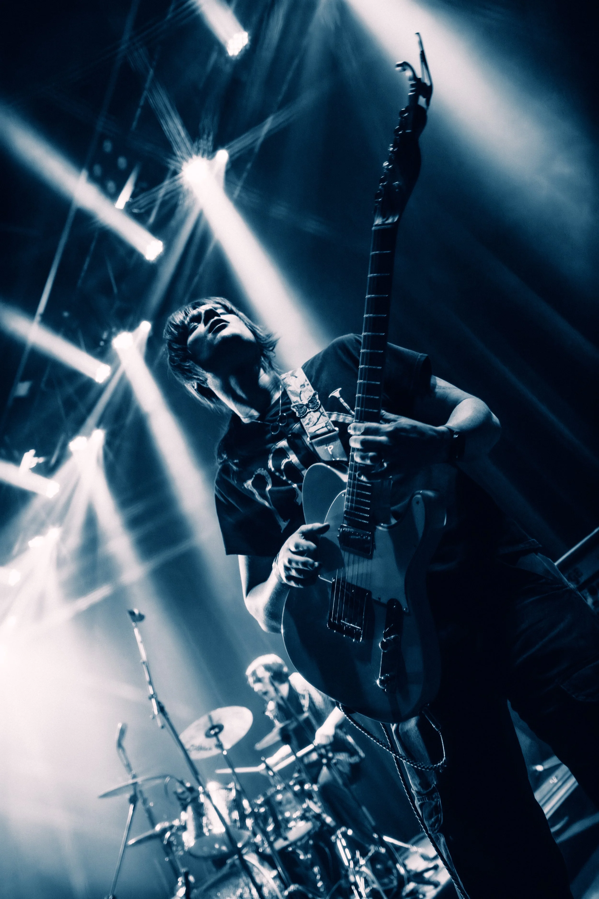 A musician playing an electric guitar on stage during a concert, with stage lights shining down, and a drummer in the background.