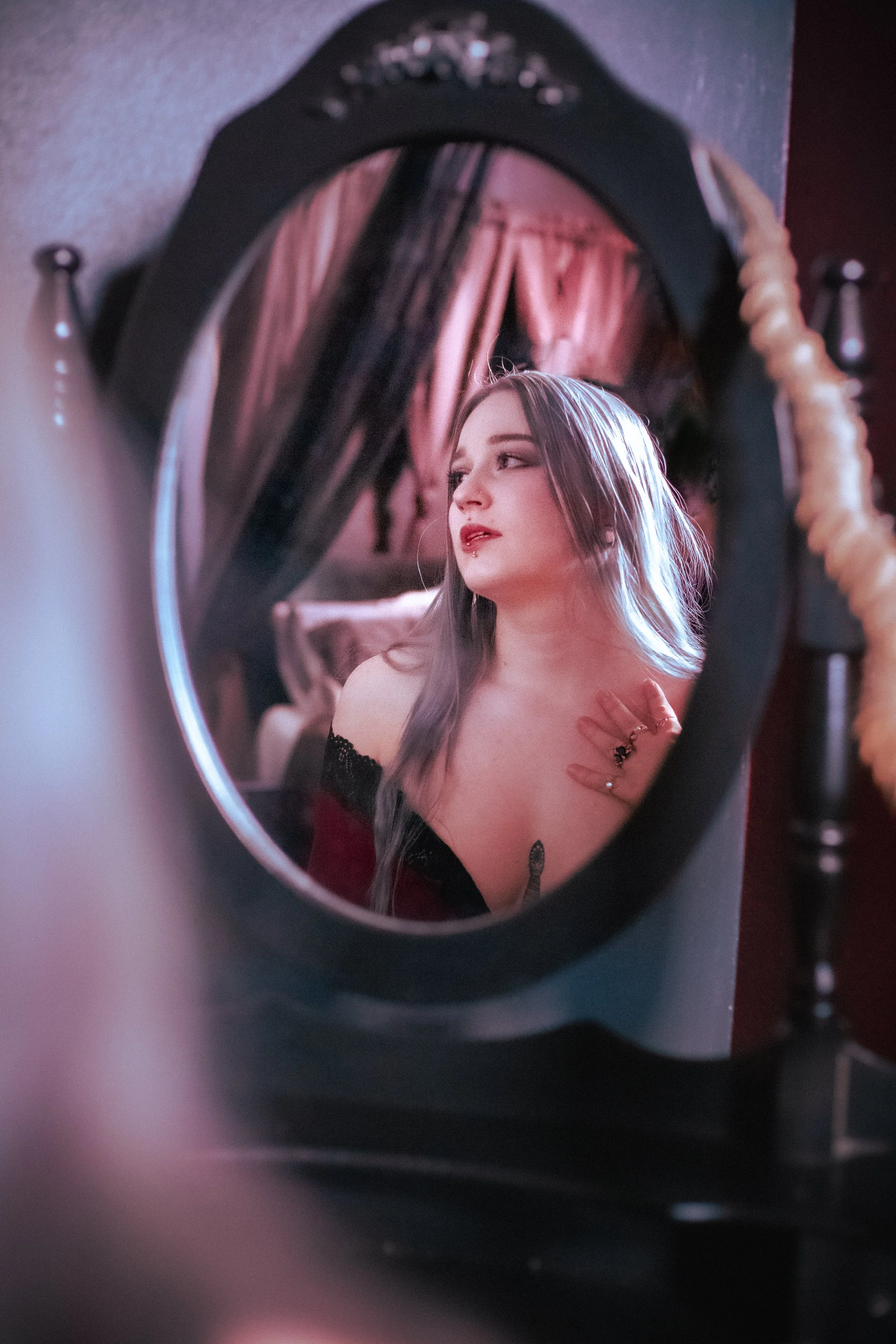 A young woman with long blonde hair looking at herself in a mirror, wearing a red and black off-shoulder top, with tattoos on her arm and shoulder, and a piercing on her lower lip.