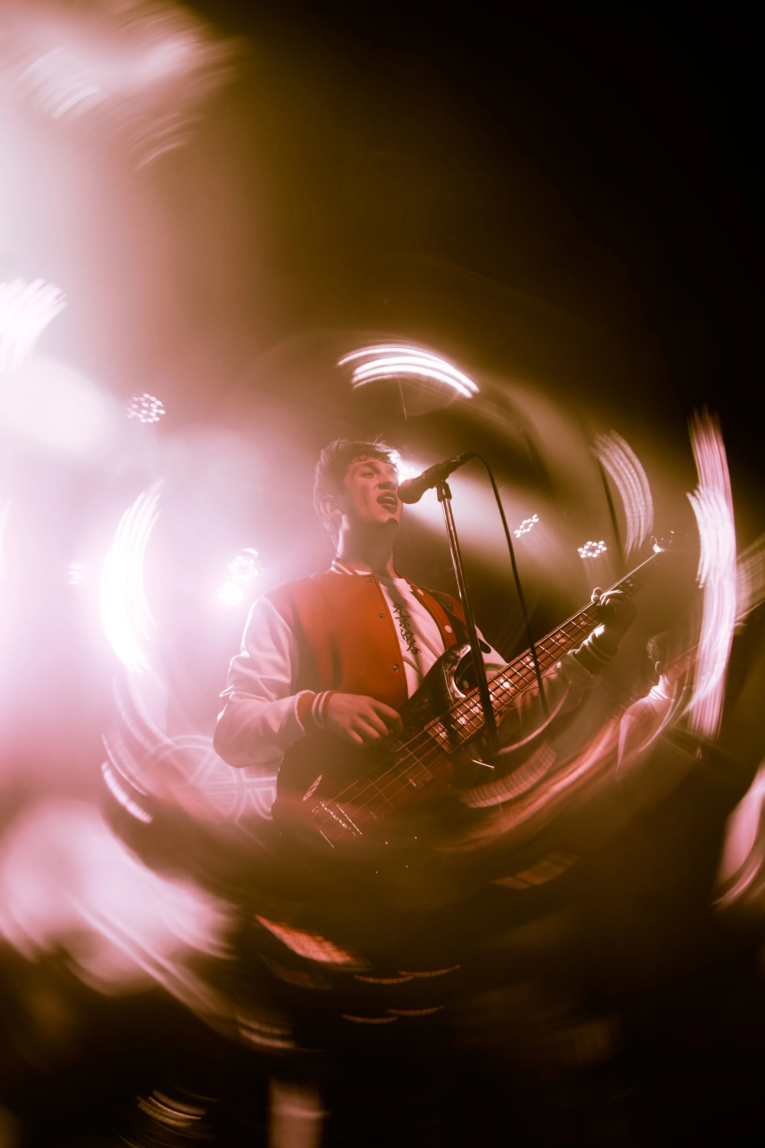 A young male musician performs on stage with a guitar, singing into a microphone, surrounded by vibrant, swirling stage lights and effects.