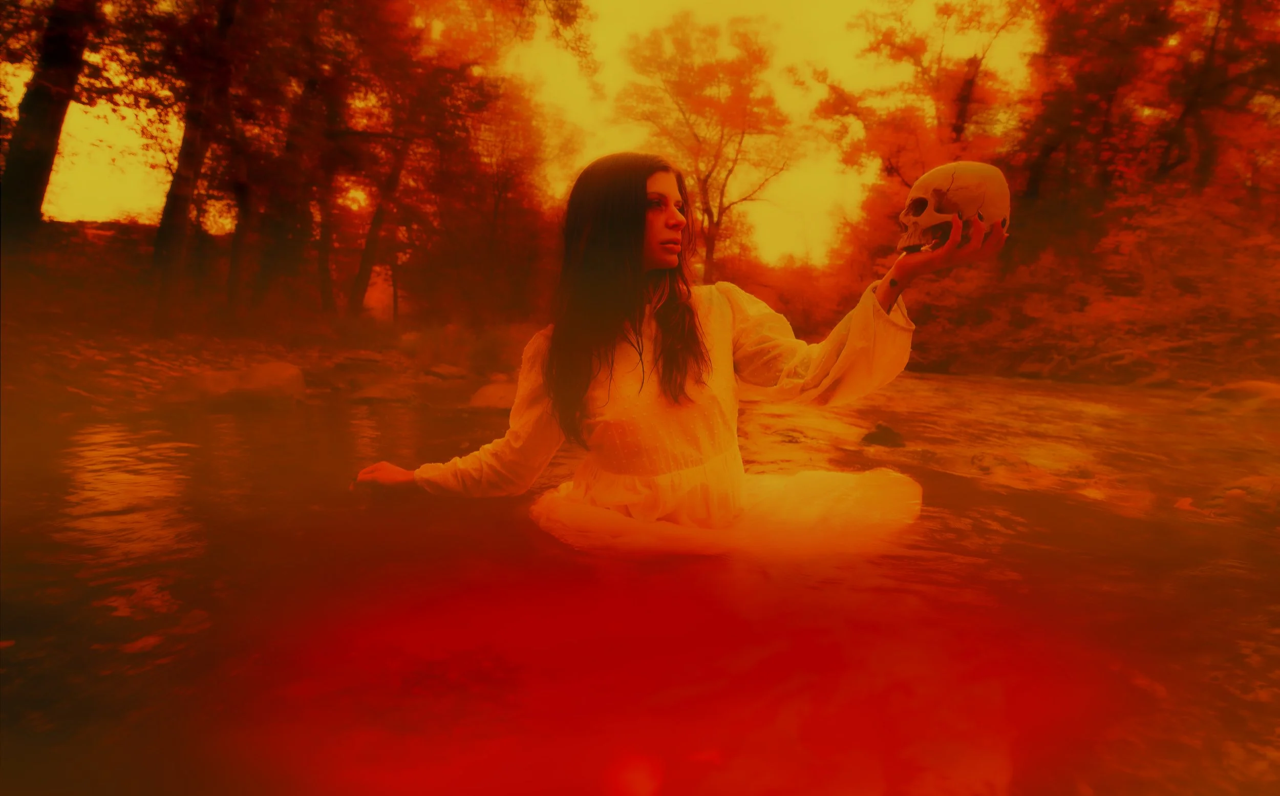 A woman with long dark hair in a yellow dress sitting in a river at sunset, holding a human skull.