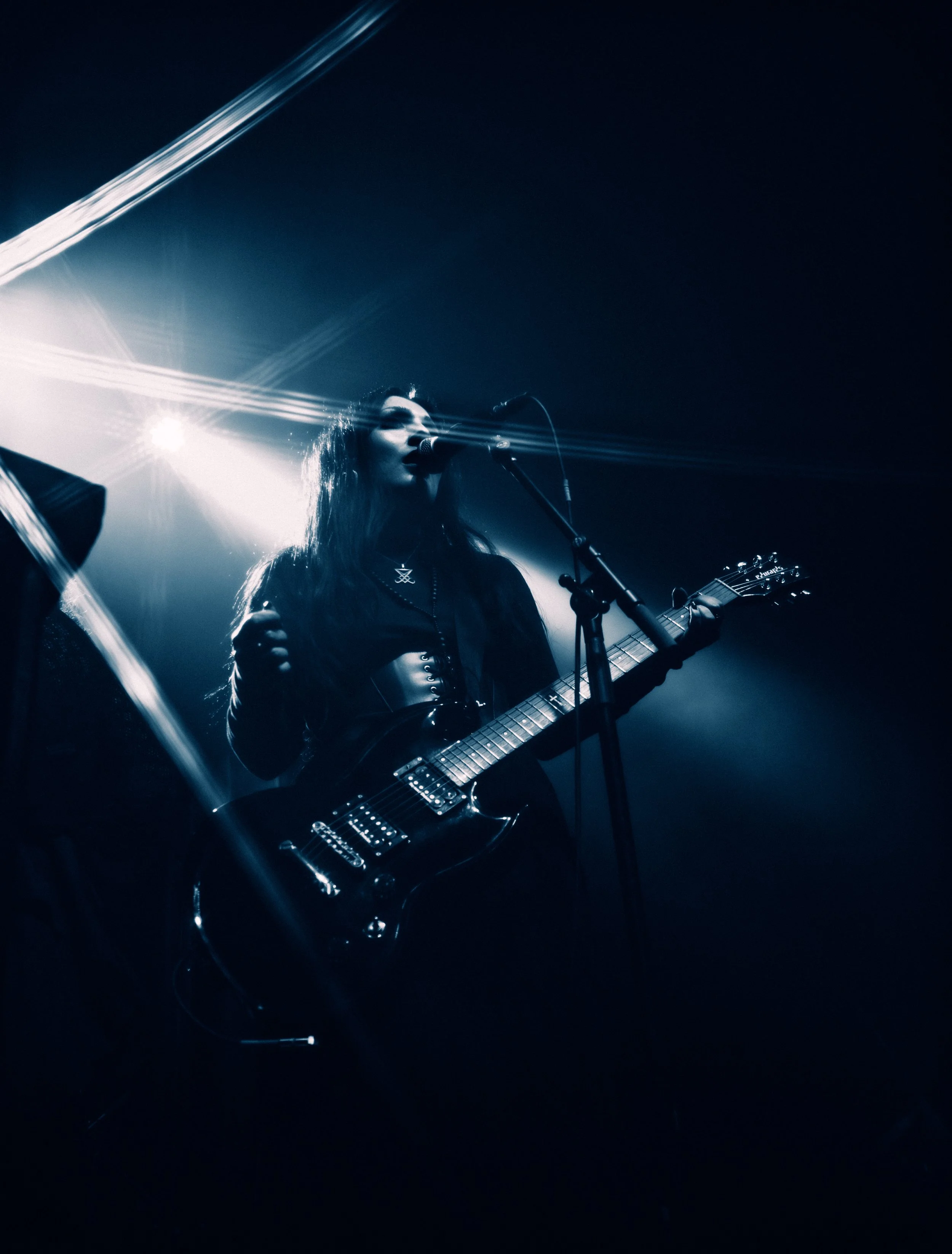 A woman playing an electric guitar and singing into a microphone on stage, with bright stage lights creating a dramatic effect.