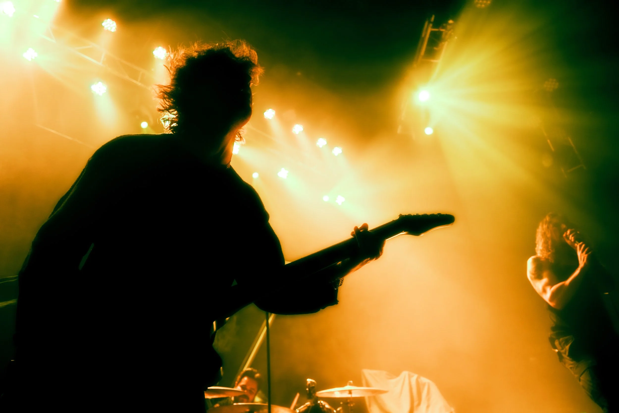 Silhouette of a guitarist on stage with bright yellow and green lights shining down, during a live concert.