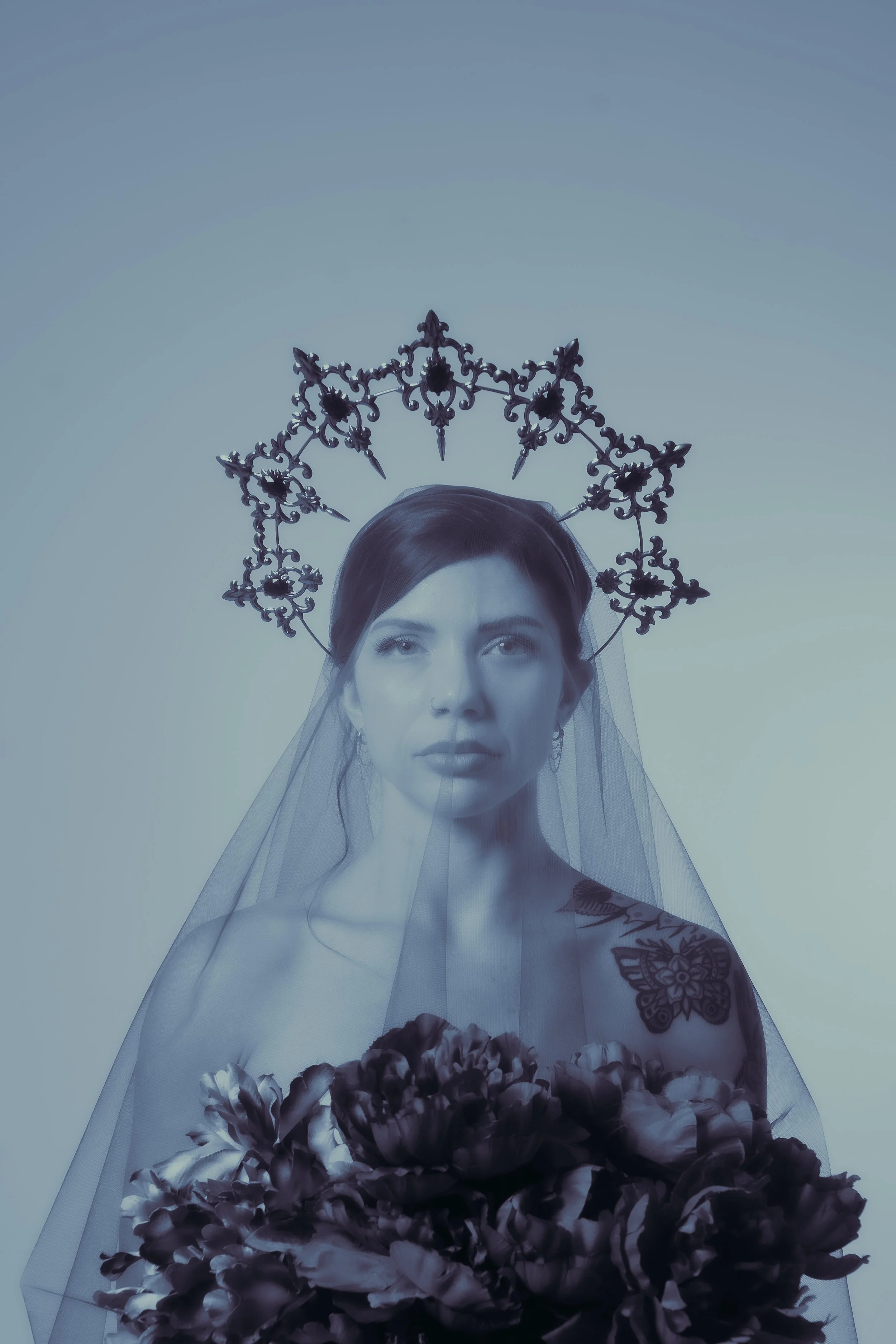 A woman wearing a veil and ornate headpiece, holding a bouquet of dark flowers, with tattoos visible on her shoulder.