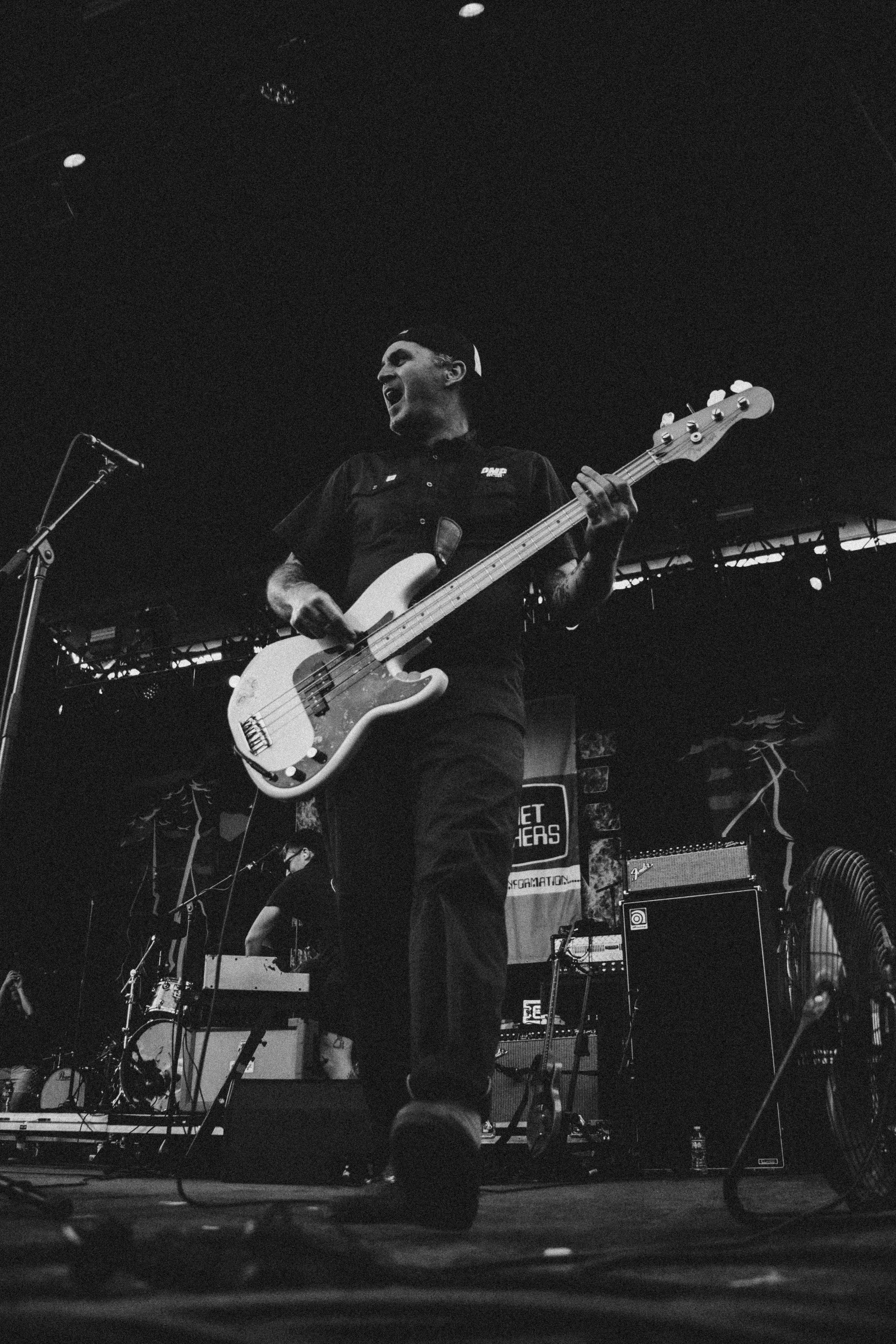 A musician passionately playing an electric bass guitar on stage during a live performance, with stage equipment and other band members visible in the background.