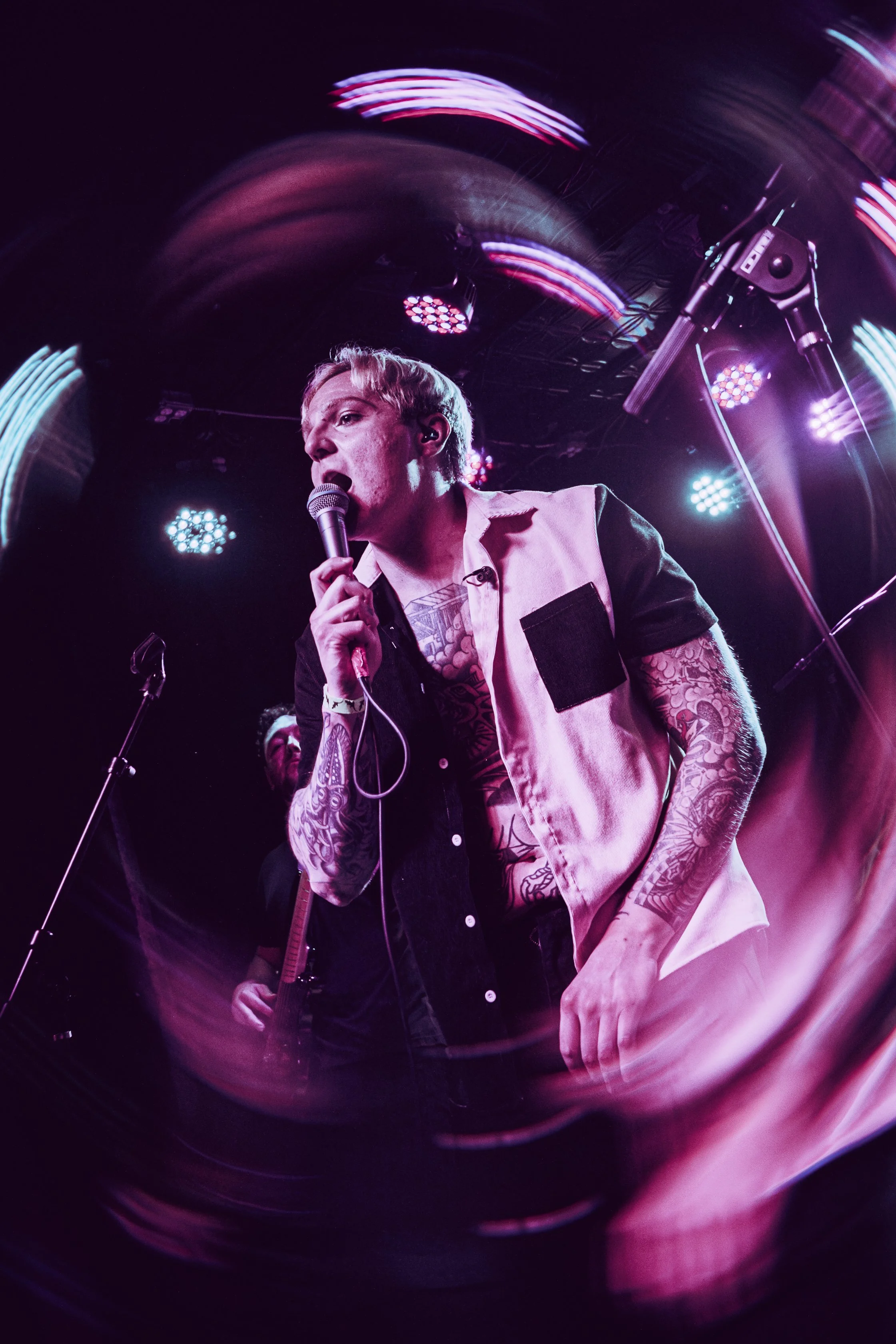 A tattooed male singer holding a microphone performs on stage with colorful lights and a band in the background.