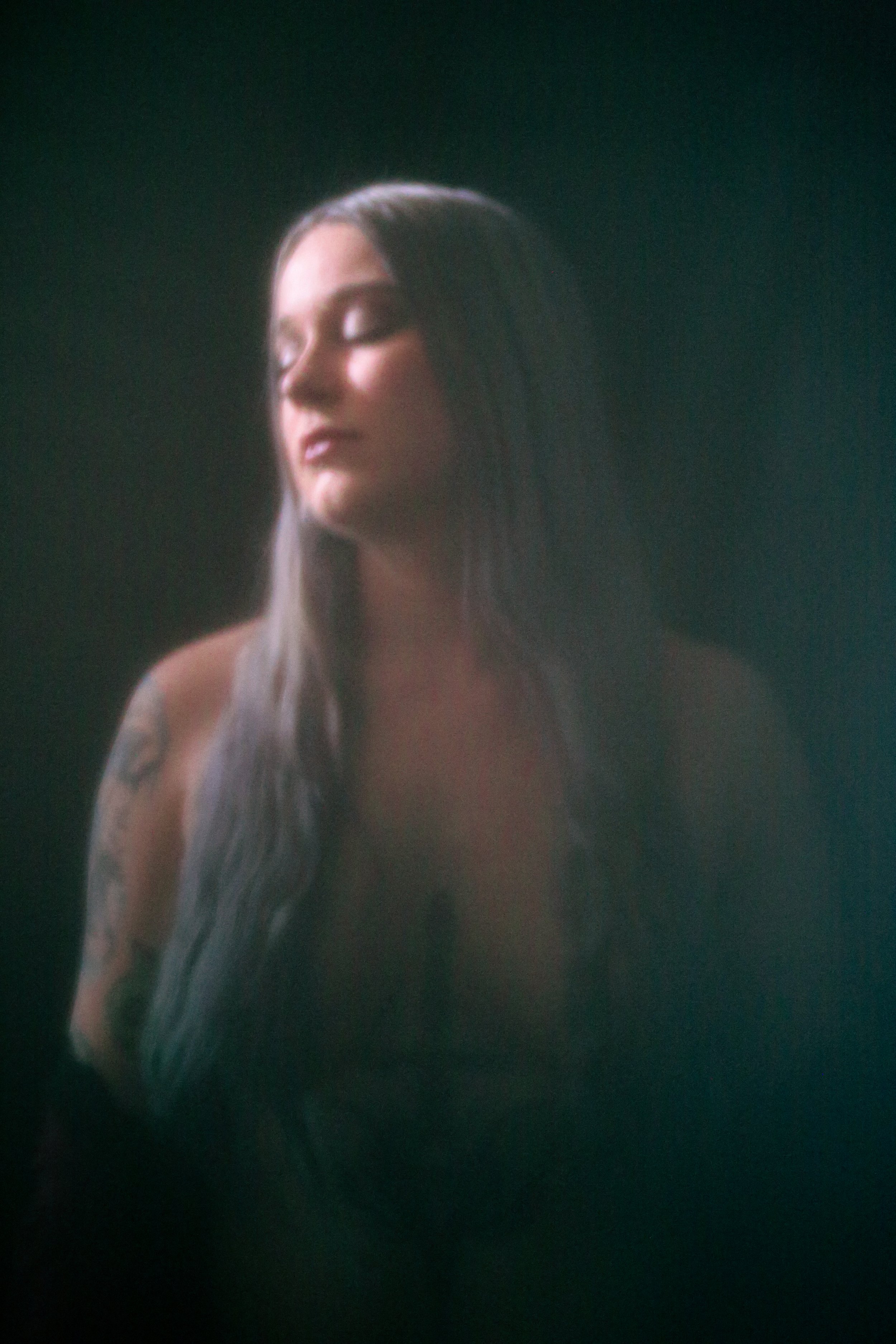 Portrait of a woman with long dark hair and tattoos on her arm, eyes closed, soft lighting on her face against a dark background.